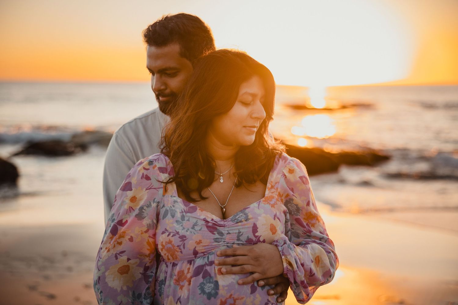 Couple embraces on beach at sunset with waves crashing on rocks in golden light as sun dips below horizon.