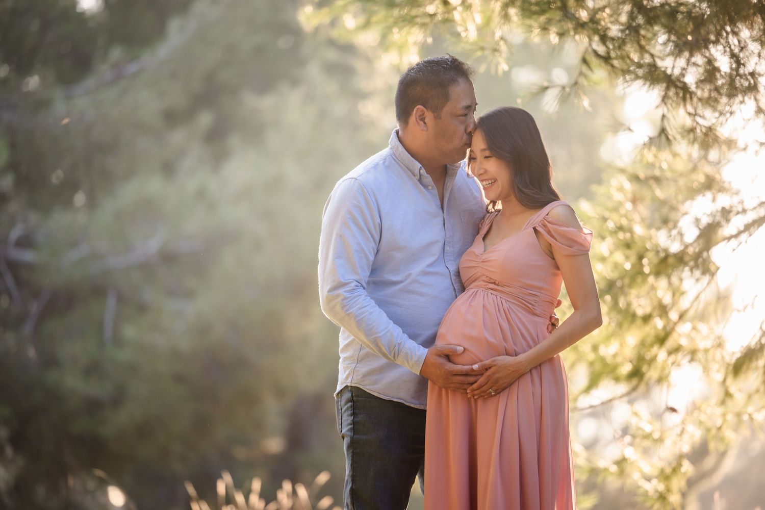 A couple shares a tender moment at sunset while expecting their first child, with the pregnant woman wearing a pink dress.