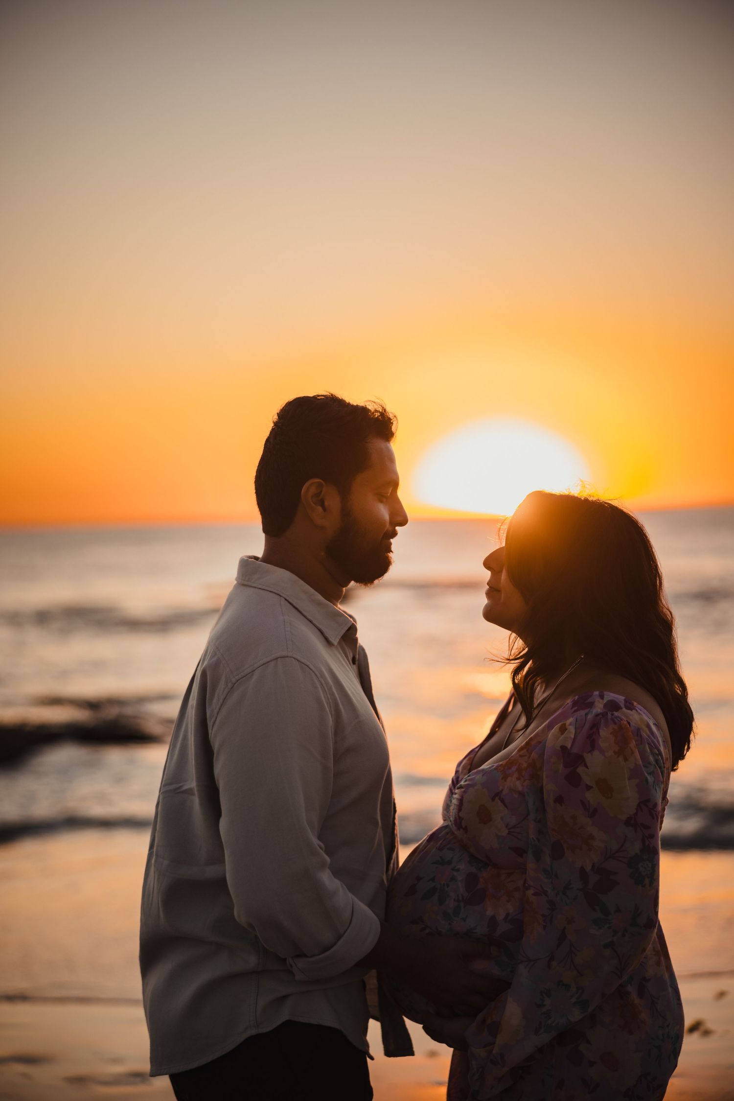 Silhouetted couple stands intimately on beach during golden hour sunset over ocean.