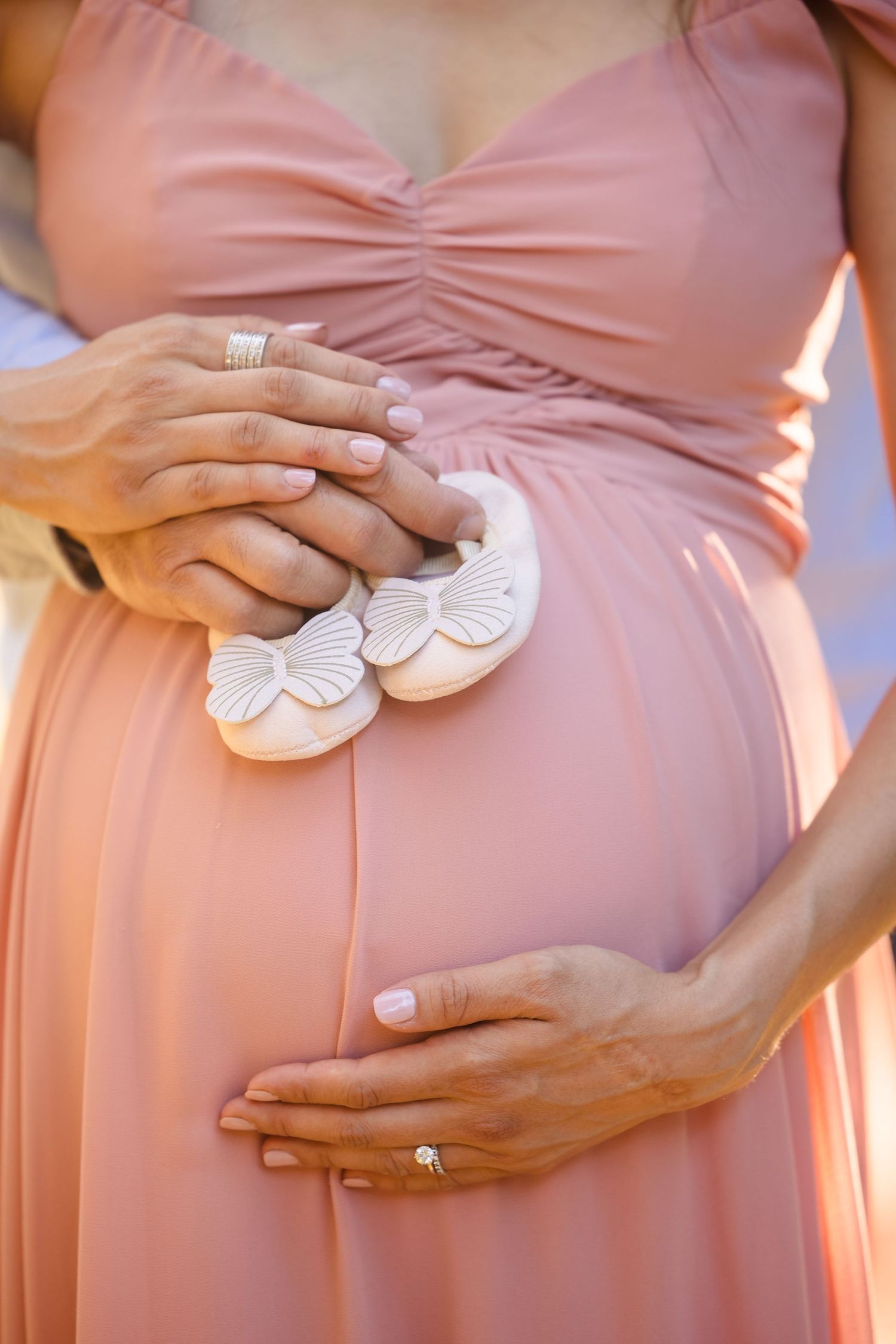 Hands holding tiny white baby shoes against a peach-colored maternity dress, showcasing a pregnancy moment.
