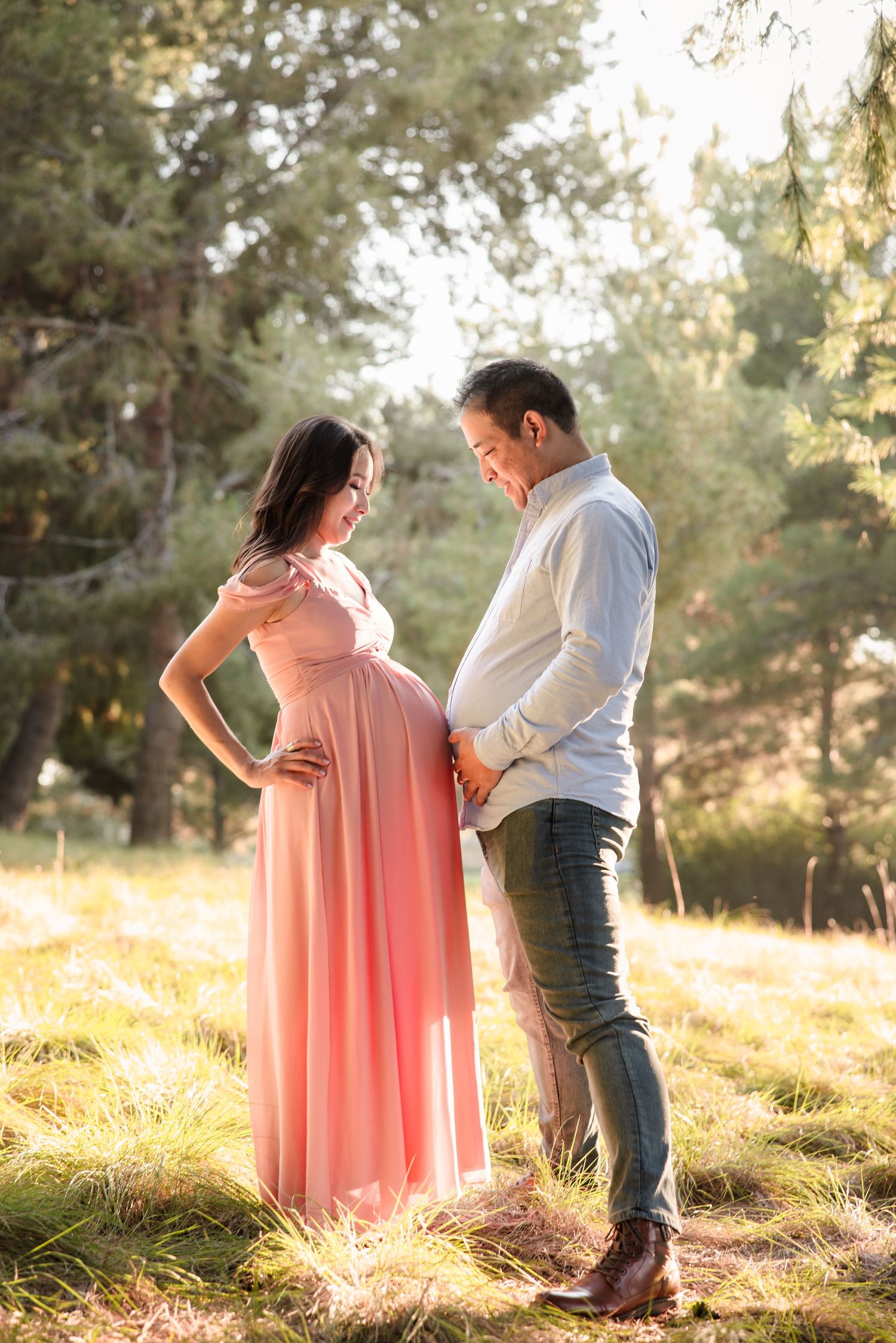 Couple standing in sunlit meadow with trees as expectant mother wears flowing coral dress for maternity photoshoot.