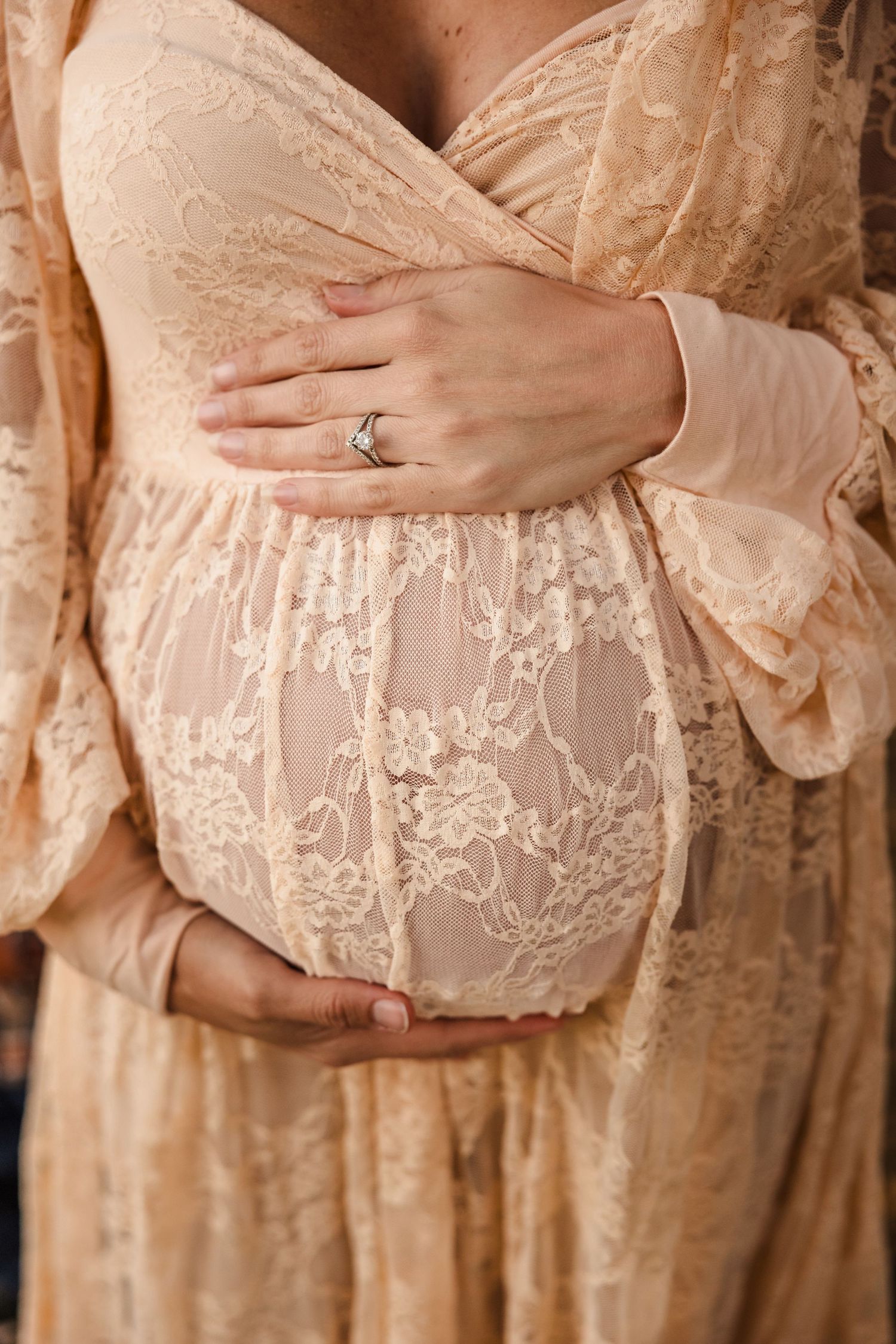 Close-up of pregnant belly wrapped in delicate beige lace dress with hands displaying engagement ring.