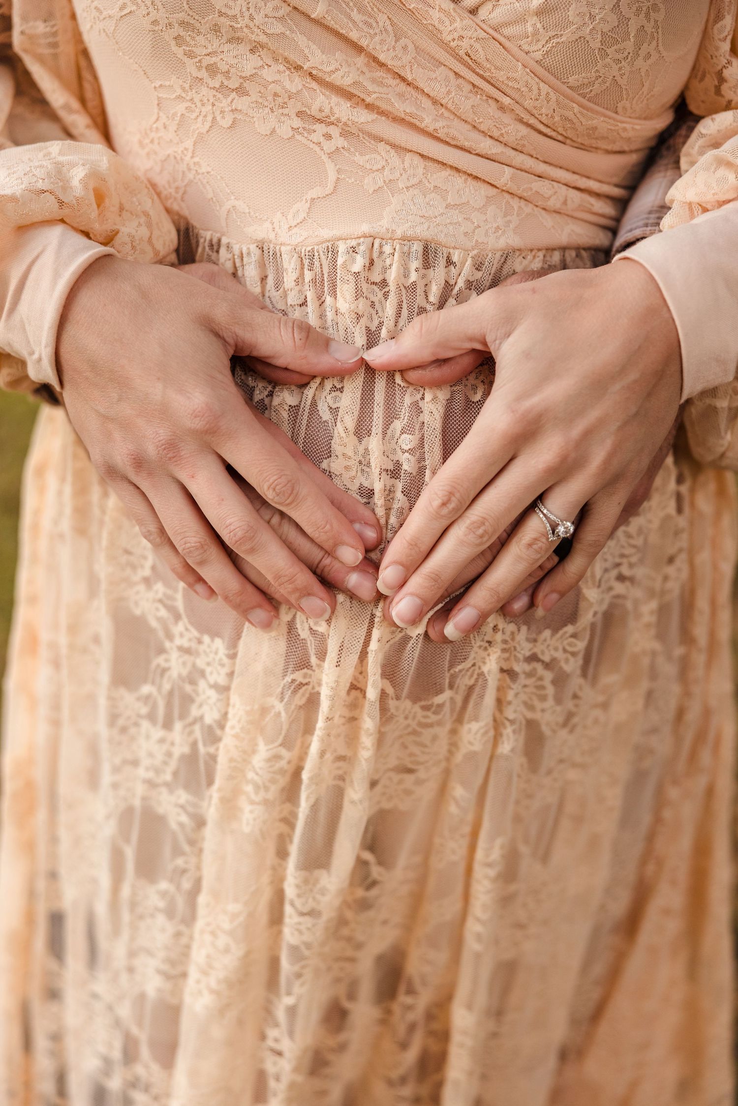 Delicate hands rest on a cream-colored lace dress, adorned with an engagement ring in soft natural lighting.