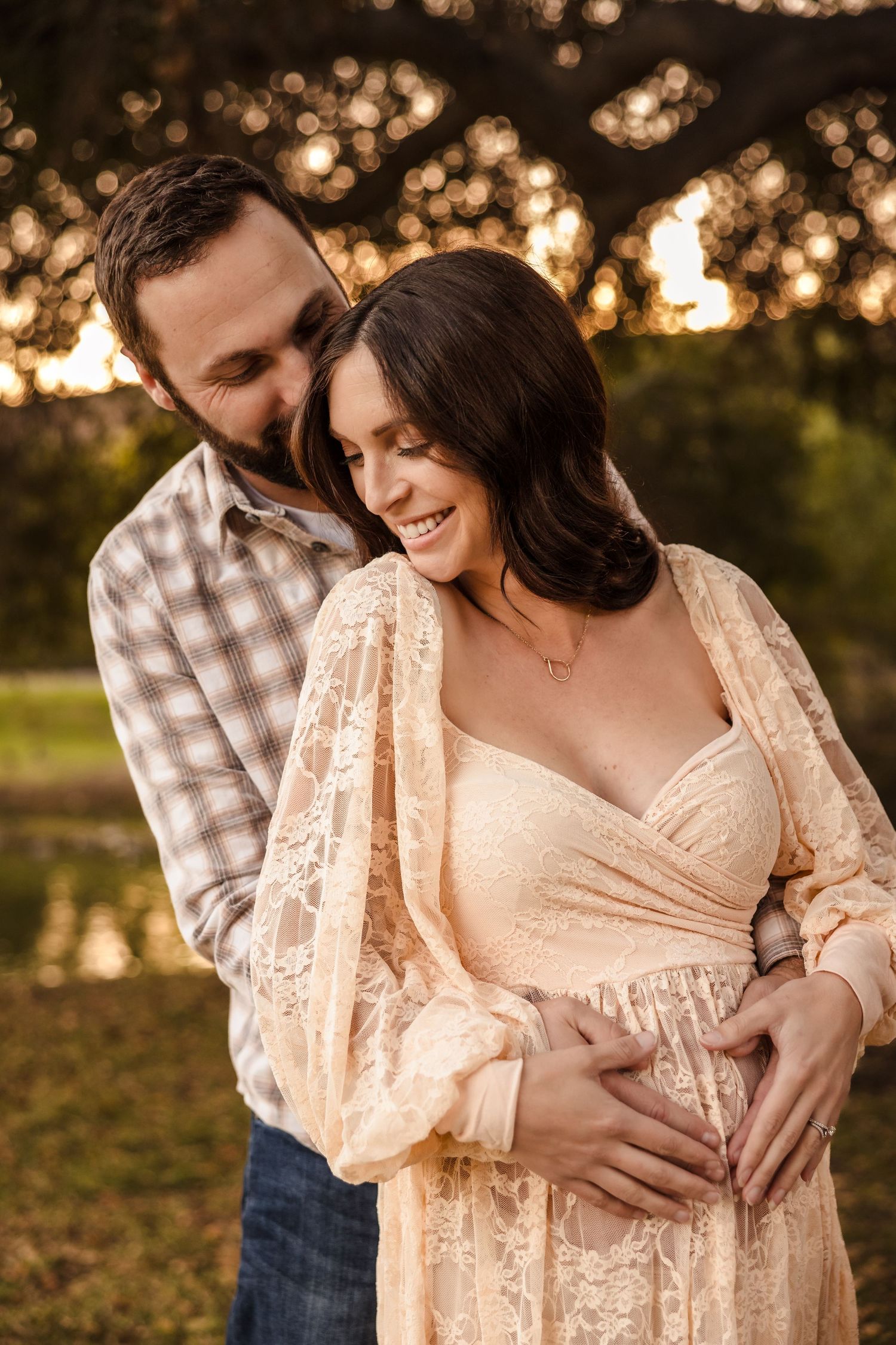 A couple embraces at sunset while celebrating a pregnancy, wearing a lace dress and plaid shirt in golden light outdoors.