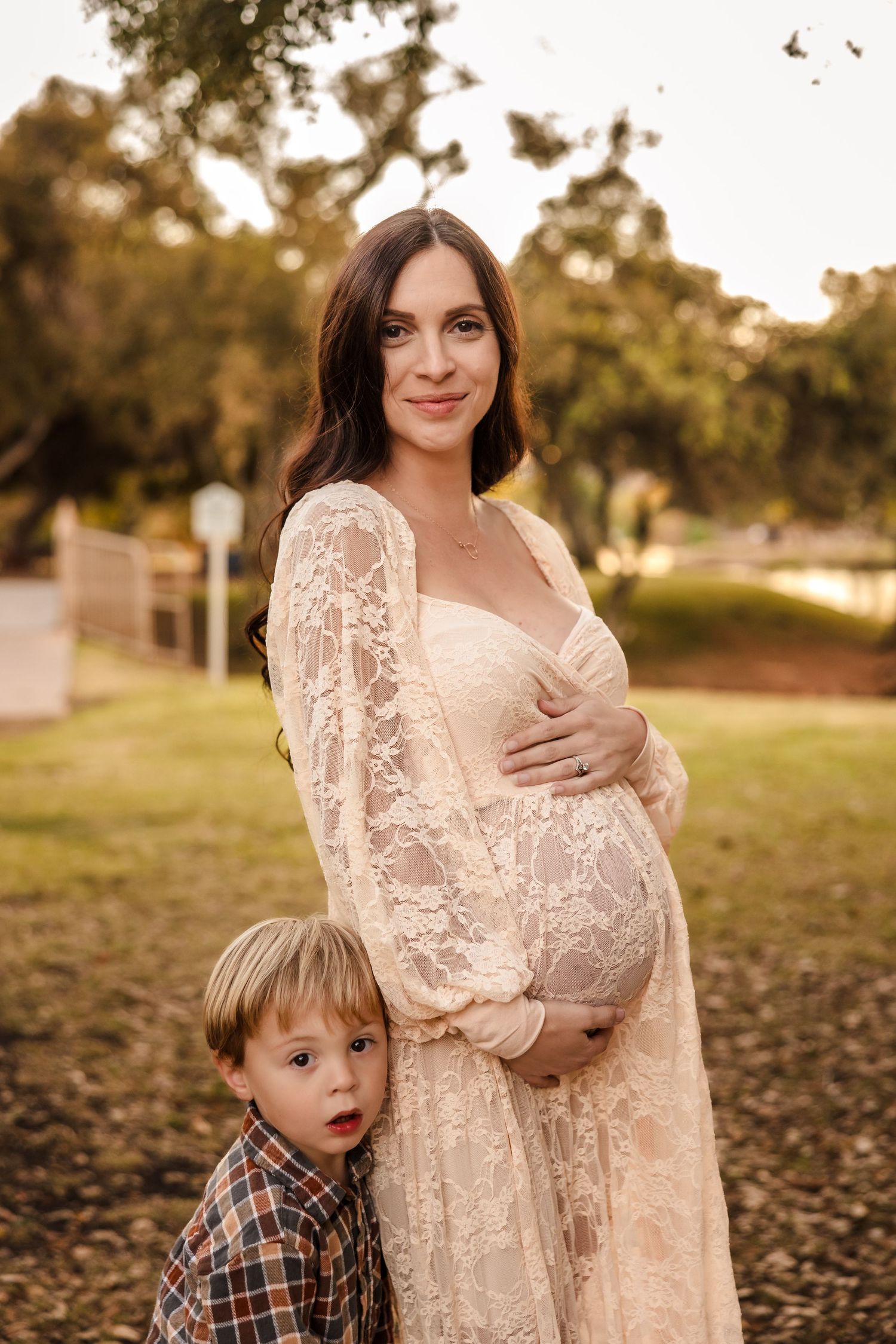 Maternity photo outdoors at sunset shows a woman in lace dress with a child in plaid beside her.