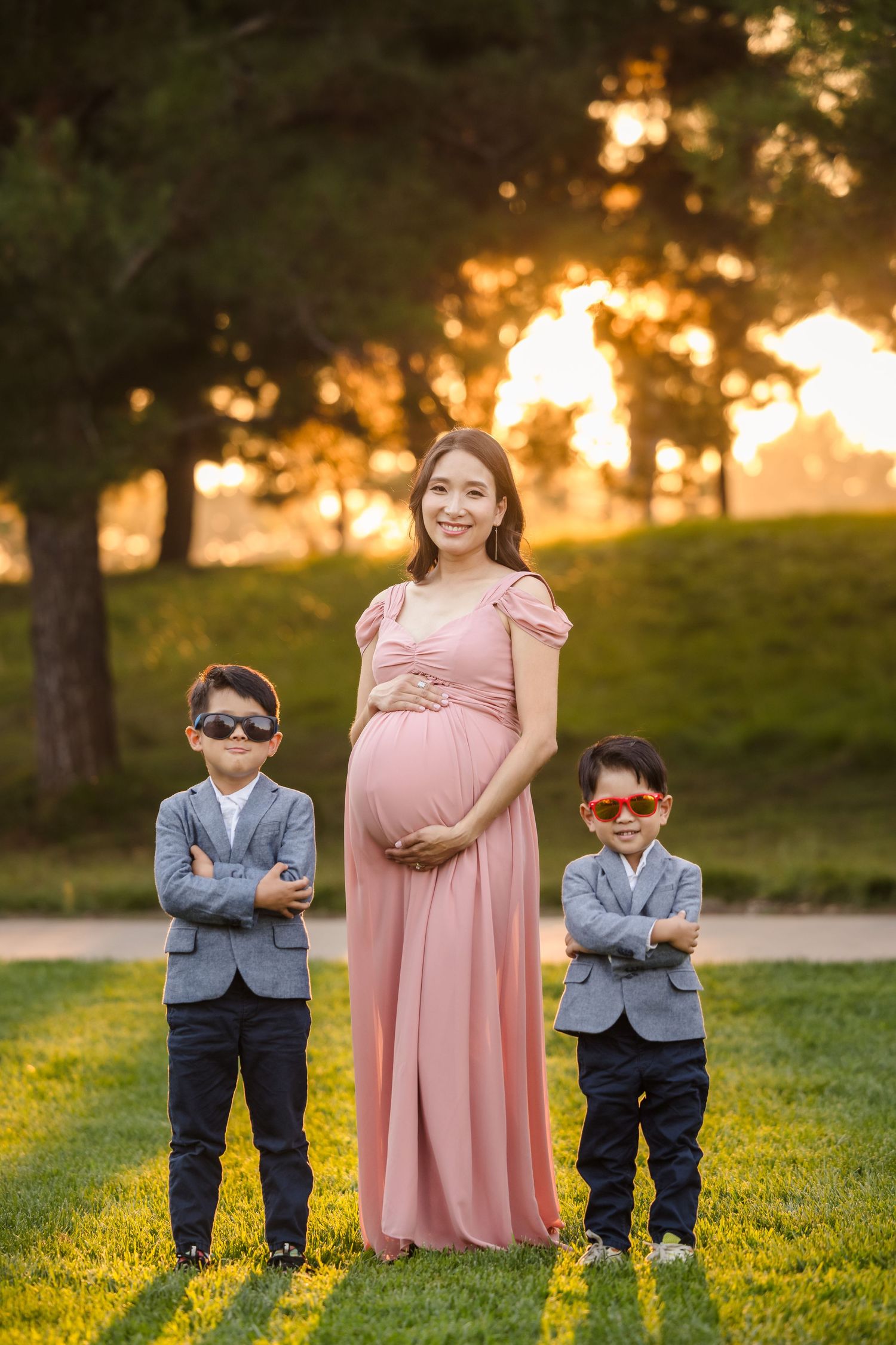 Three figures pose in a park during golden hour sunset, with young boys in suits and sunglasses beside a woman in pink dress.
