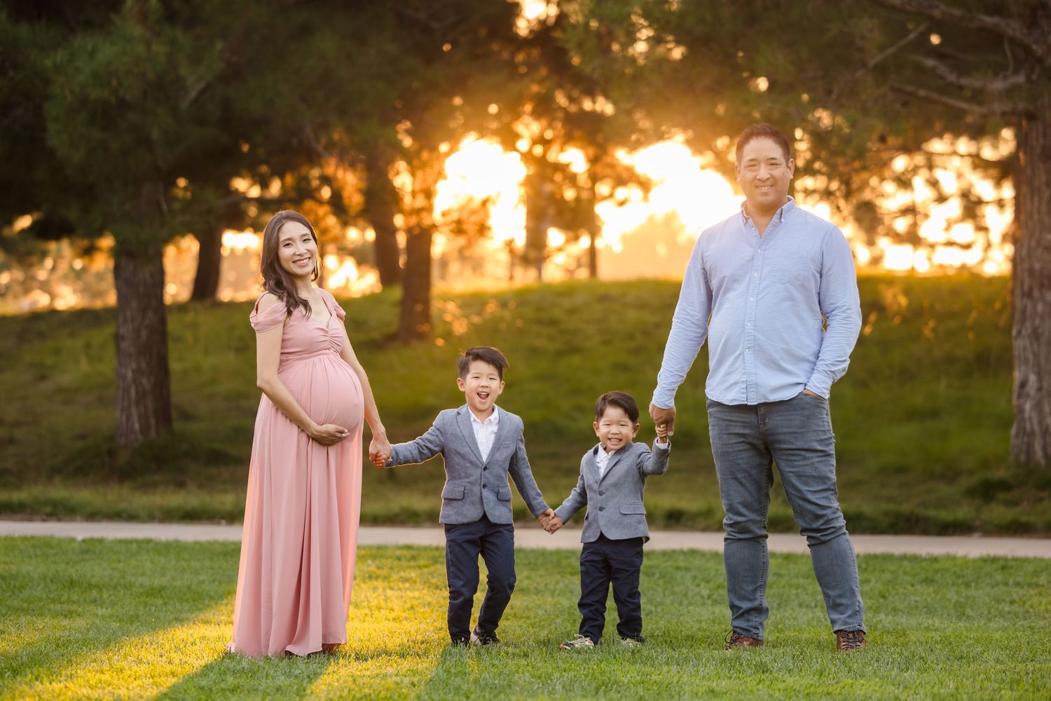 Family poses together in park during golden sunset with children holding hands between parents on grassy lawn.