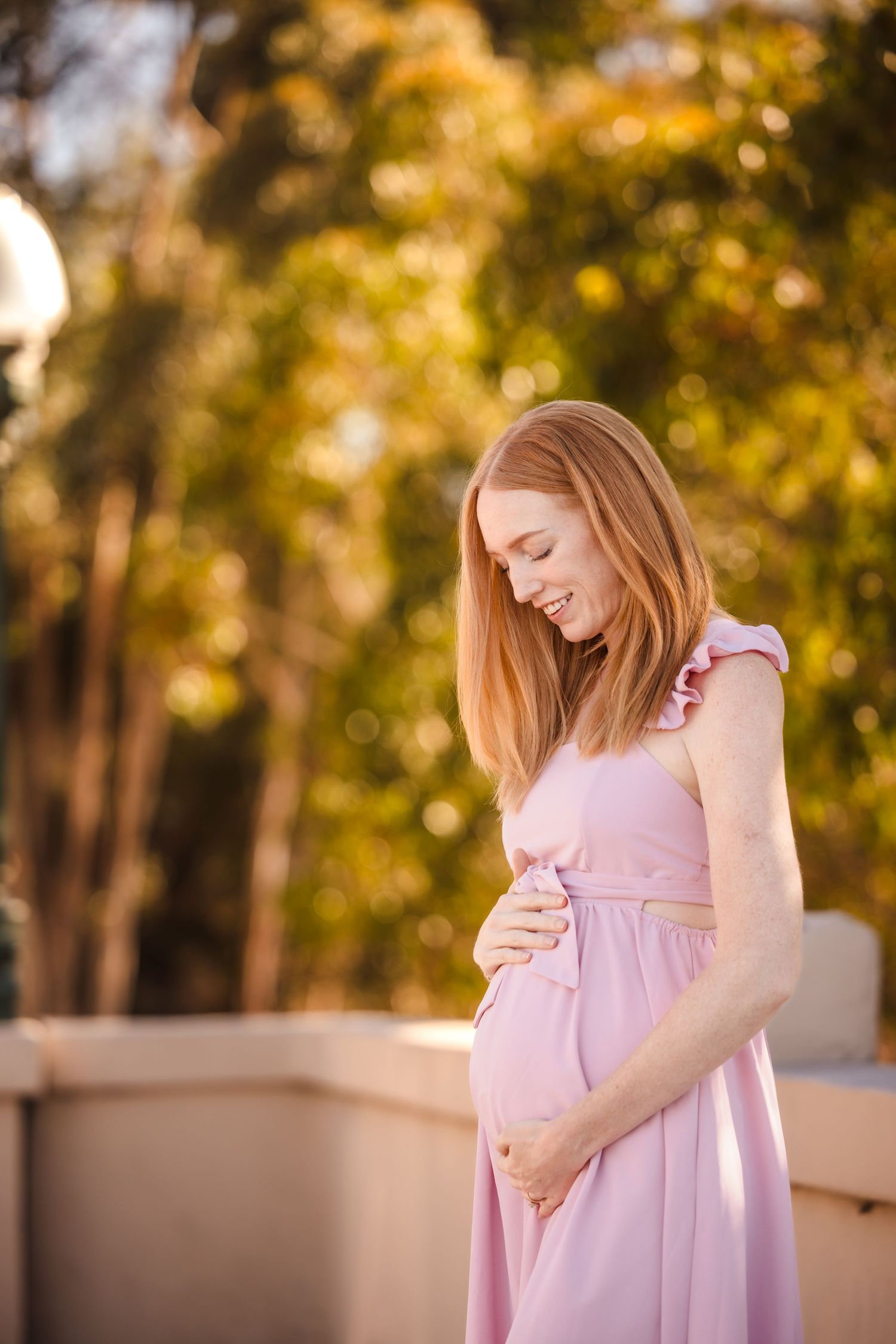 A woman in a flowing pink maternity dress lovingly cradles her baby bump in a warm autumn setting.