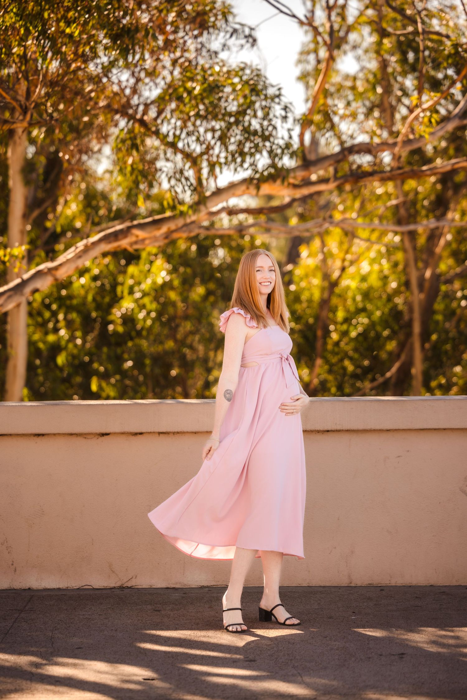 Someone in a flowing pink midi dress poses outdoors against a backdrop of sunlit trees on a warm afternoon.