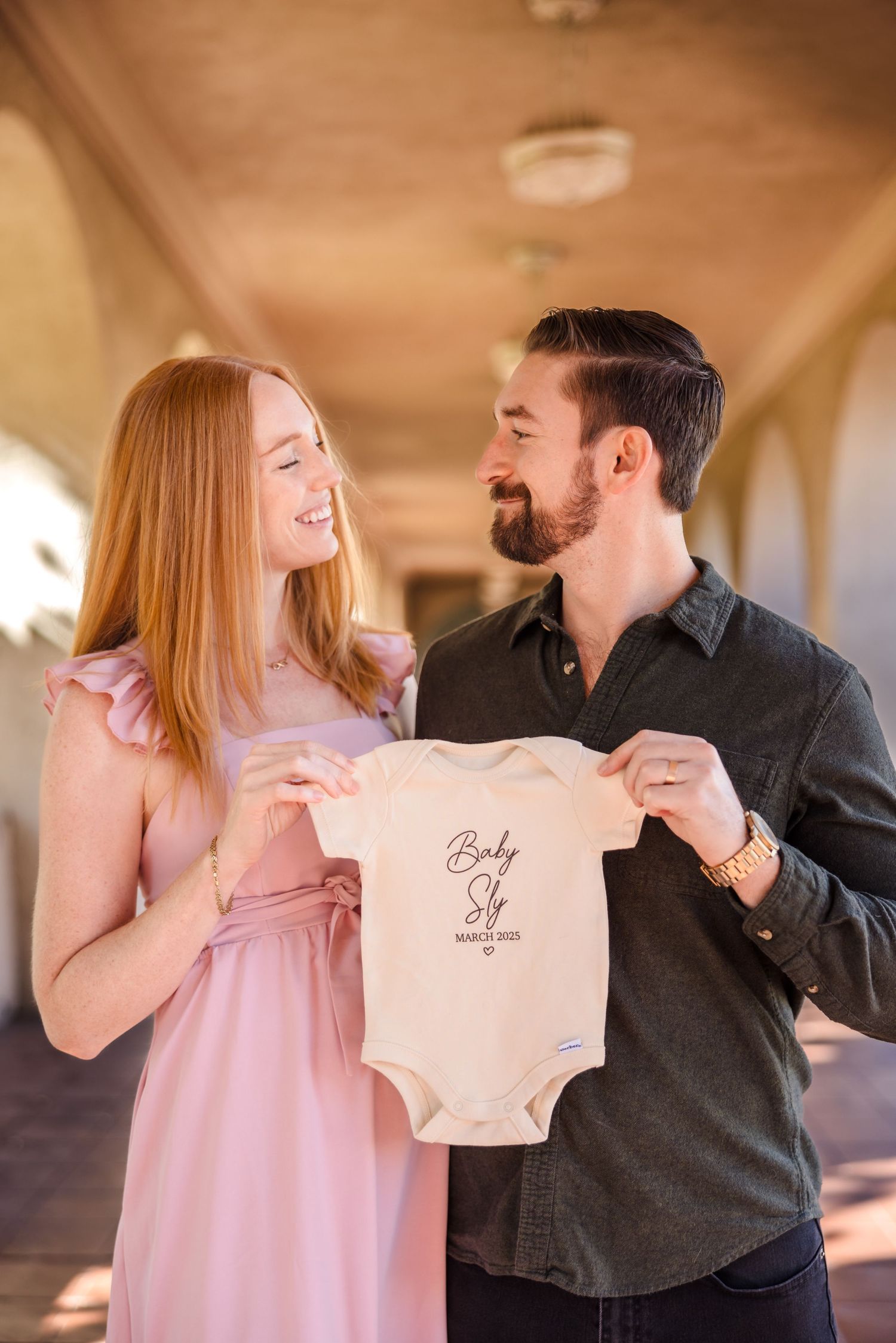 A couple stands in an arched hallway smiling while holding up a small baby onesie in warm lighting.