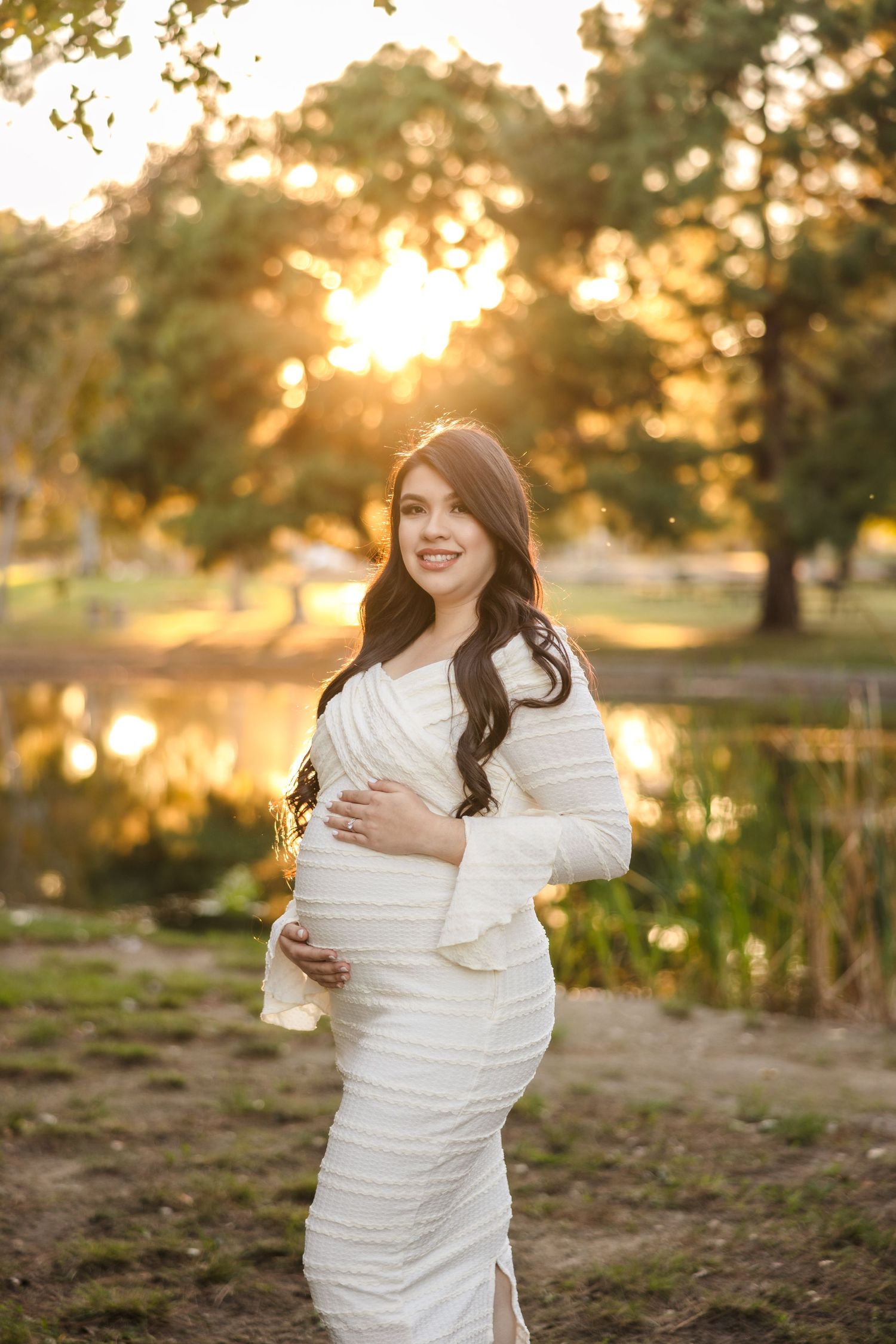 A maternity photo taken at golden hour by a pond showing a joyful moment in a white flowing dress.