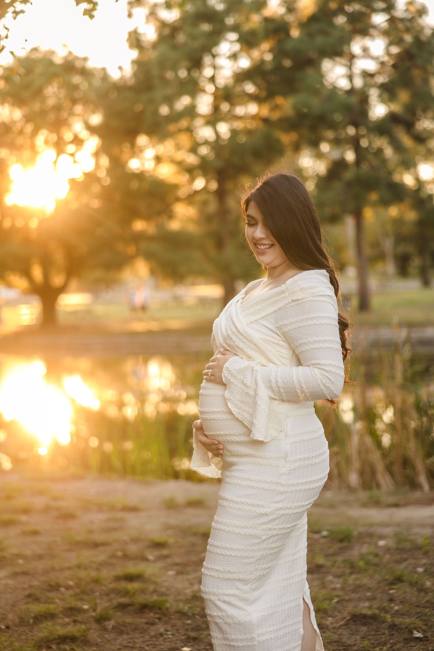 Expectant mother in white dress poses by lake at sunset with golden light filtering through trees behind her.