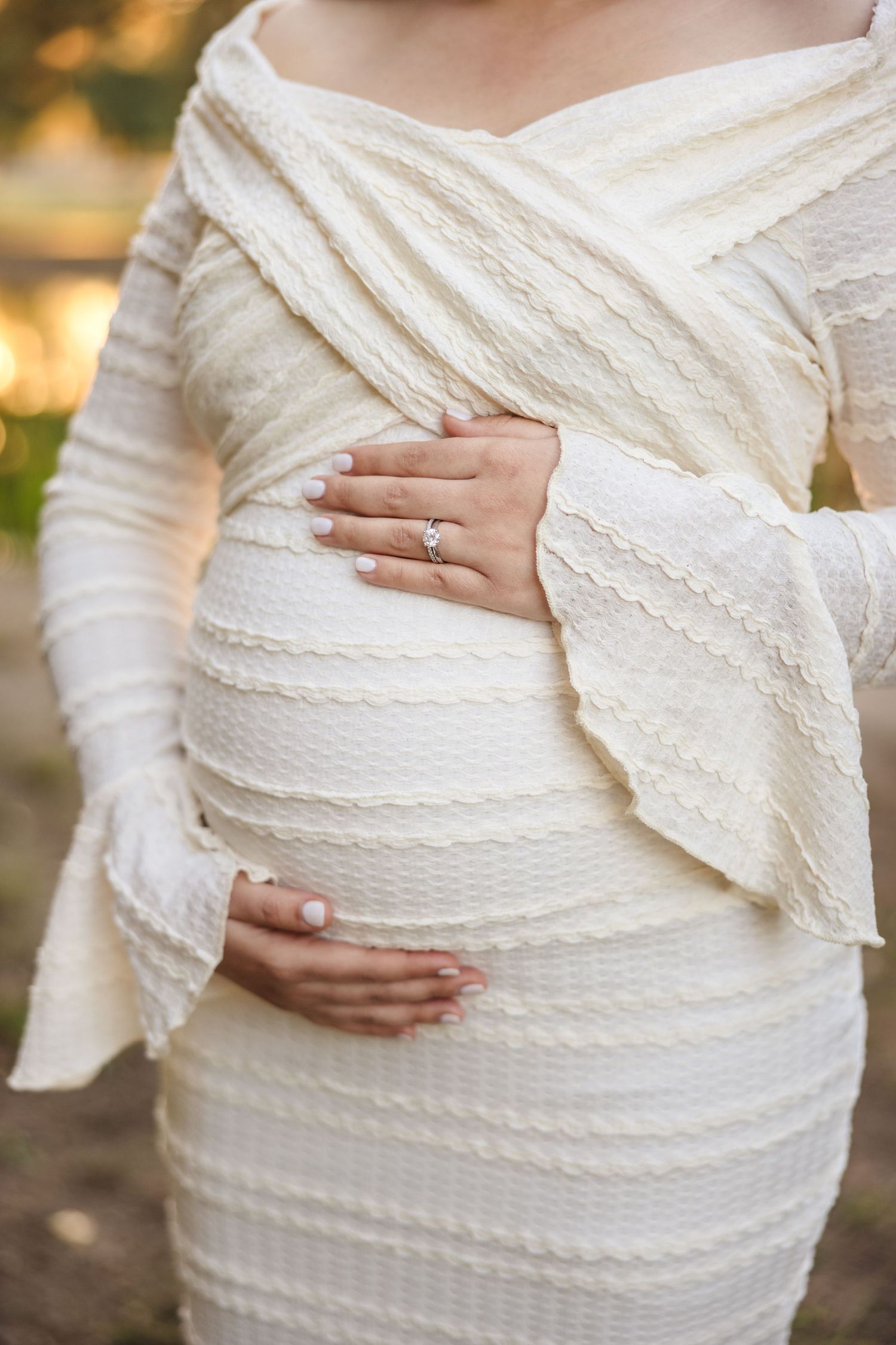 Close-up of pregnant woman wearing white lace wrap dress with hands cradling baby bump in outdoor setting.