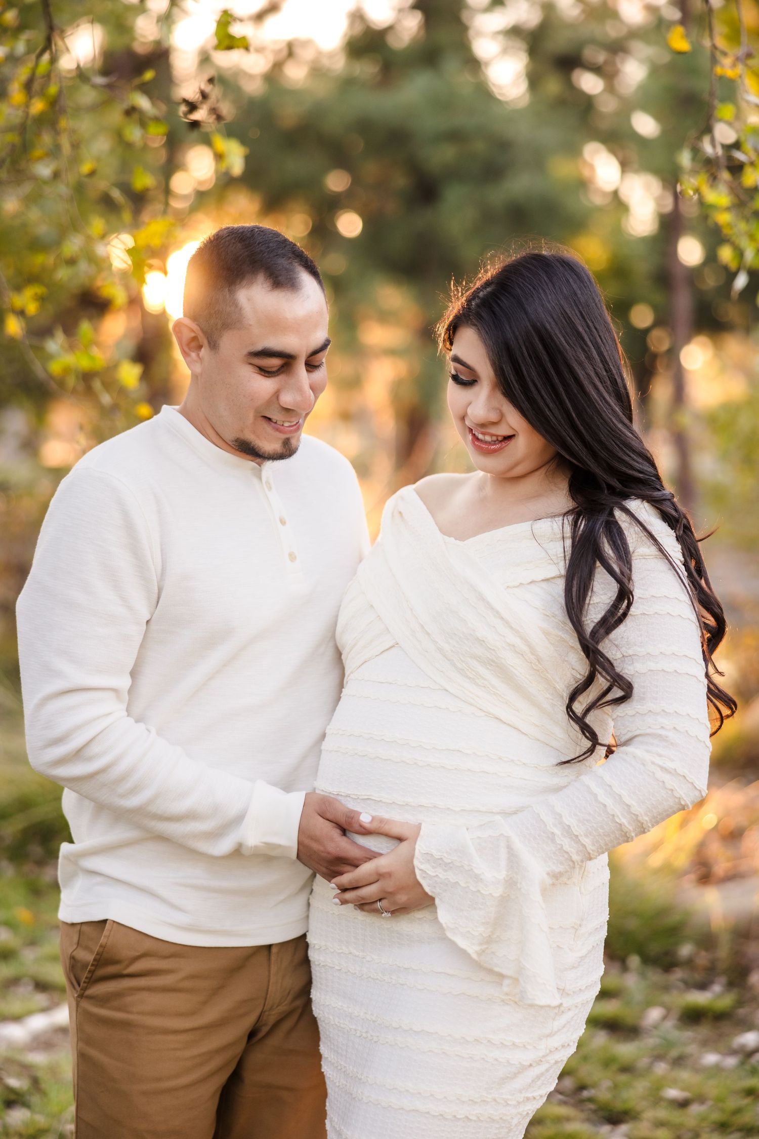 A couple in white outfits share an intimate moment during a maternity photoshoot at sunset in a garden setting.