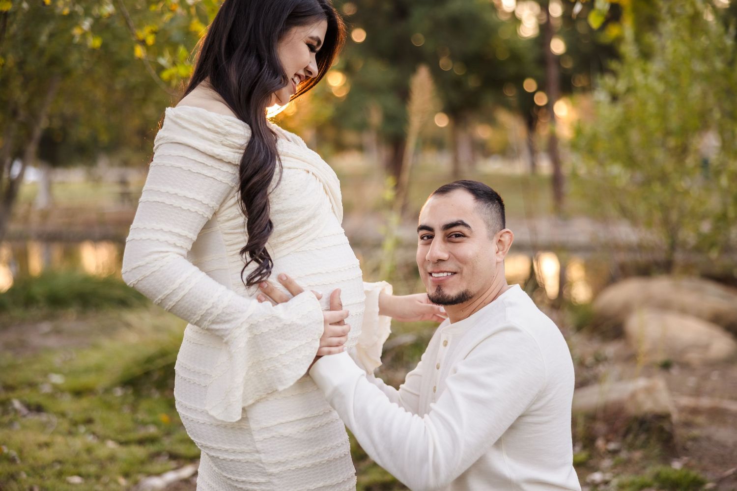 Sweet maternity photo taken outdoors with a couple in white clothing during golden hour in a natural setting.