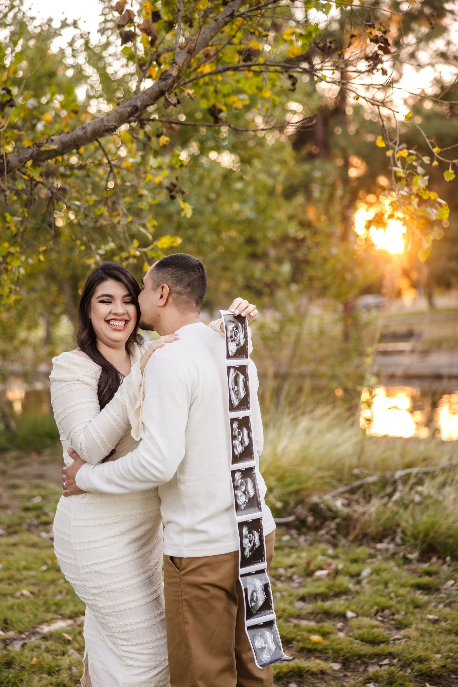 A couple shares a tender moment at sunset holding ultrasound photos in a natural outdoor setting.