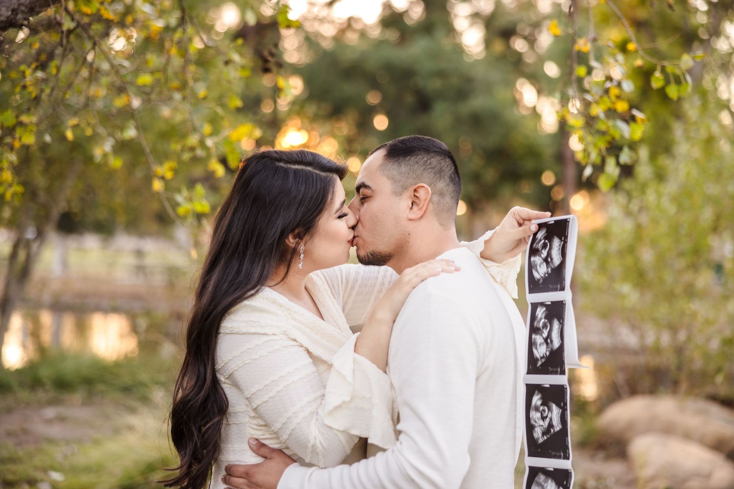 Couple in white sweaters sharing romantic moment outdoors with ultrasound photos hanging beside them.