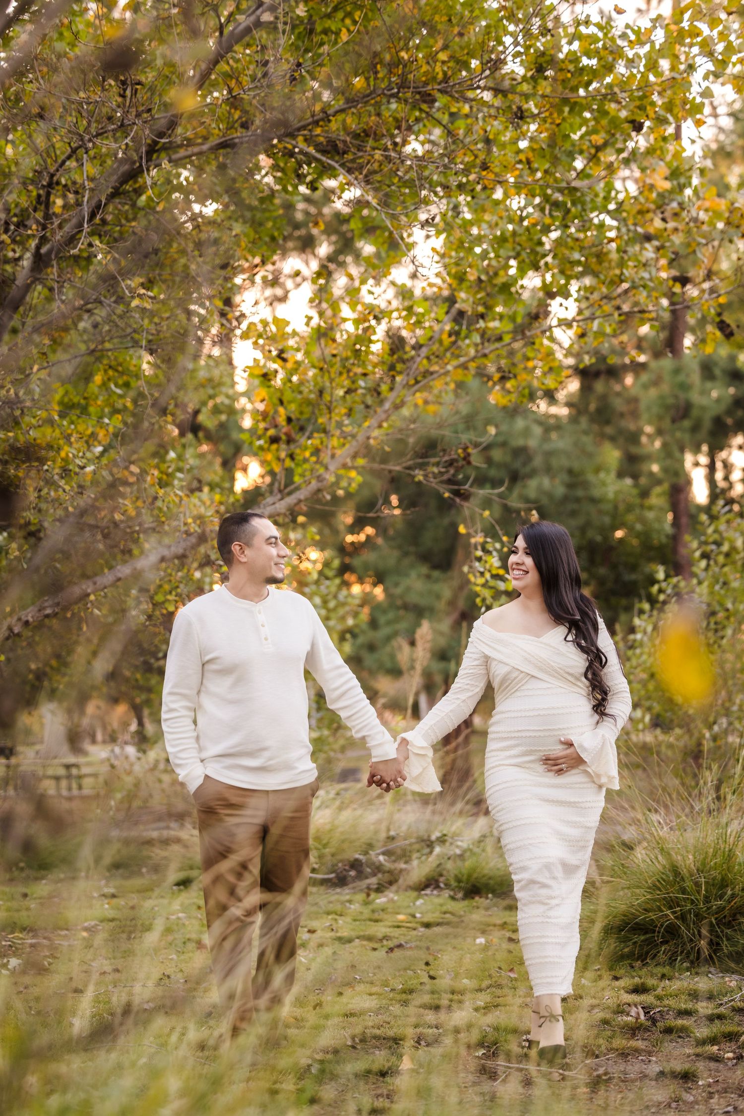 A couple in matching white outfits holds hands while walking through an autumn field at sunset.