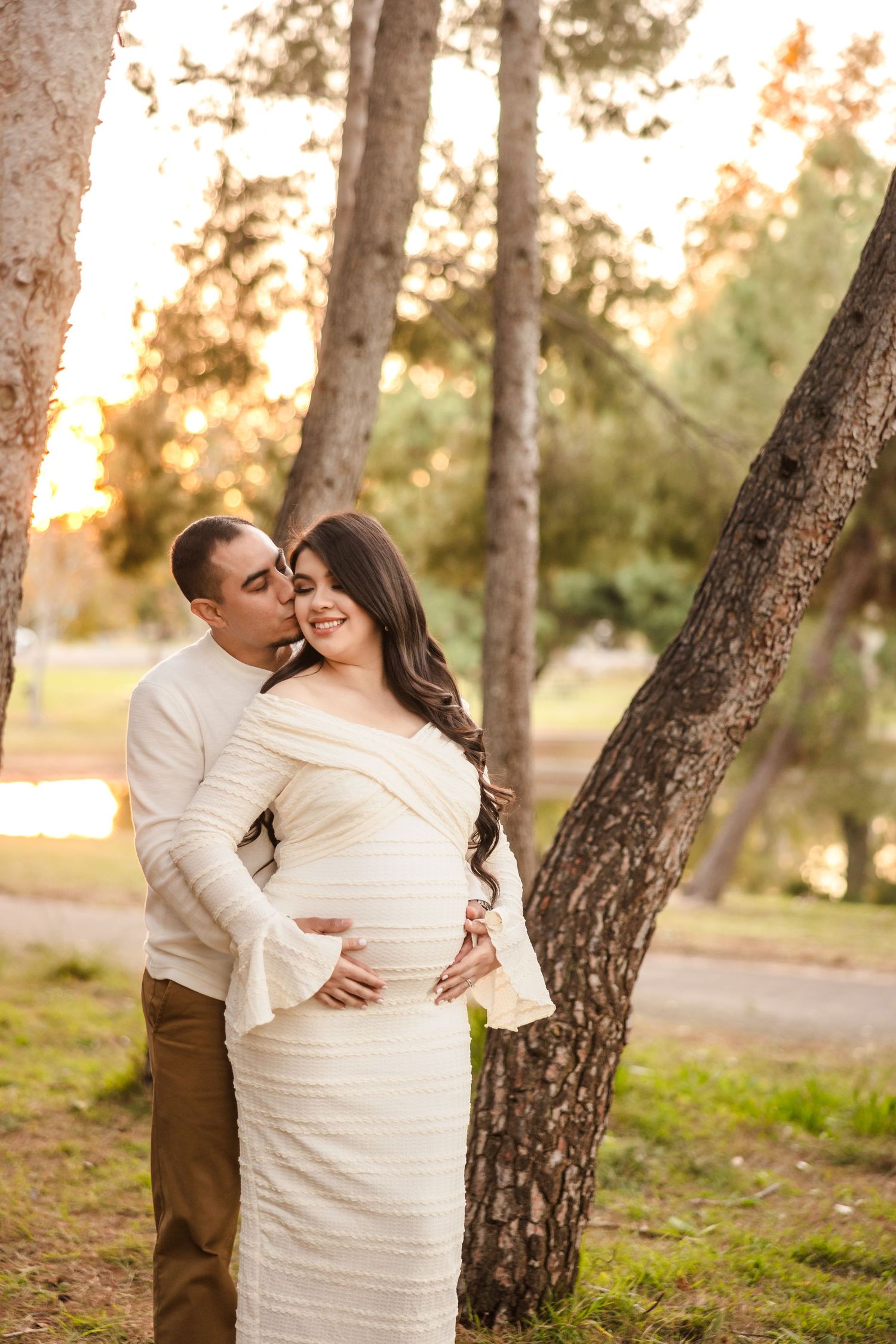 A couple embraces outdoors in a romantic woodland setting during golden hour sunset, with the woman wearing a white dress.