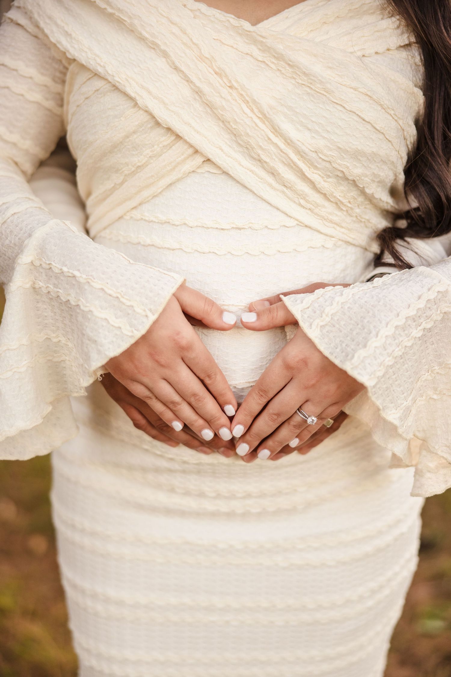Close-up of hands forming a heart shape on a pregnant belly while wearing an elegant cream-colored dress with lace details.