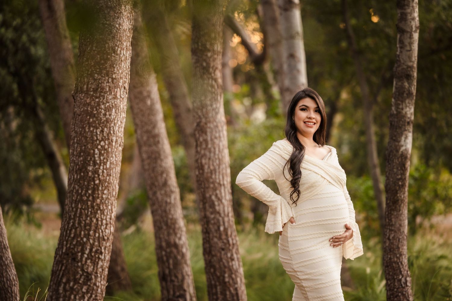 A woman wearing a cream maternity dress poses between tall pine trees during a golden hour photoshoot.