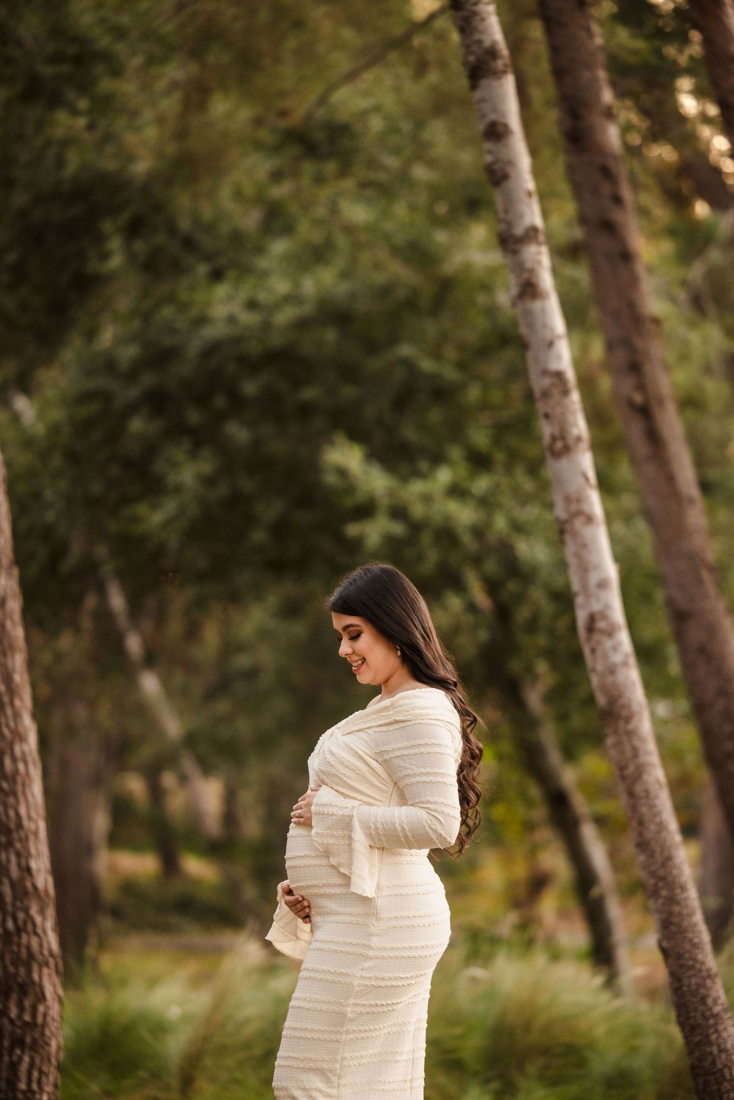 A person in a white knit dress stands in a forest setting among birch trees during golden hour.