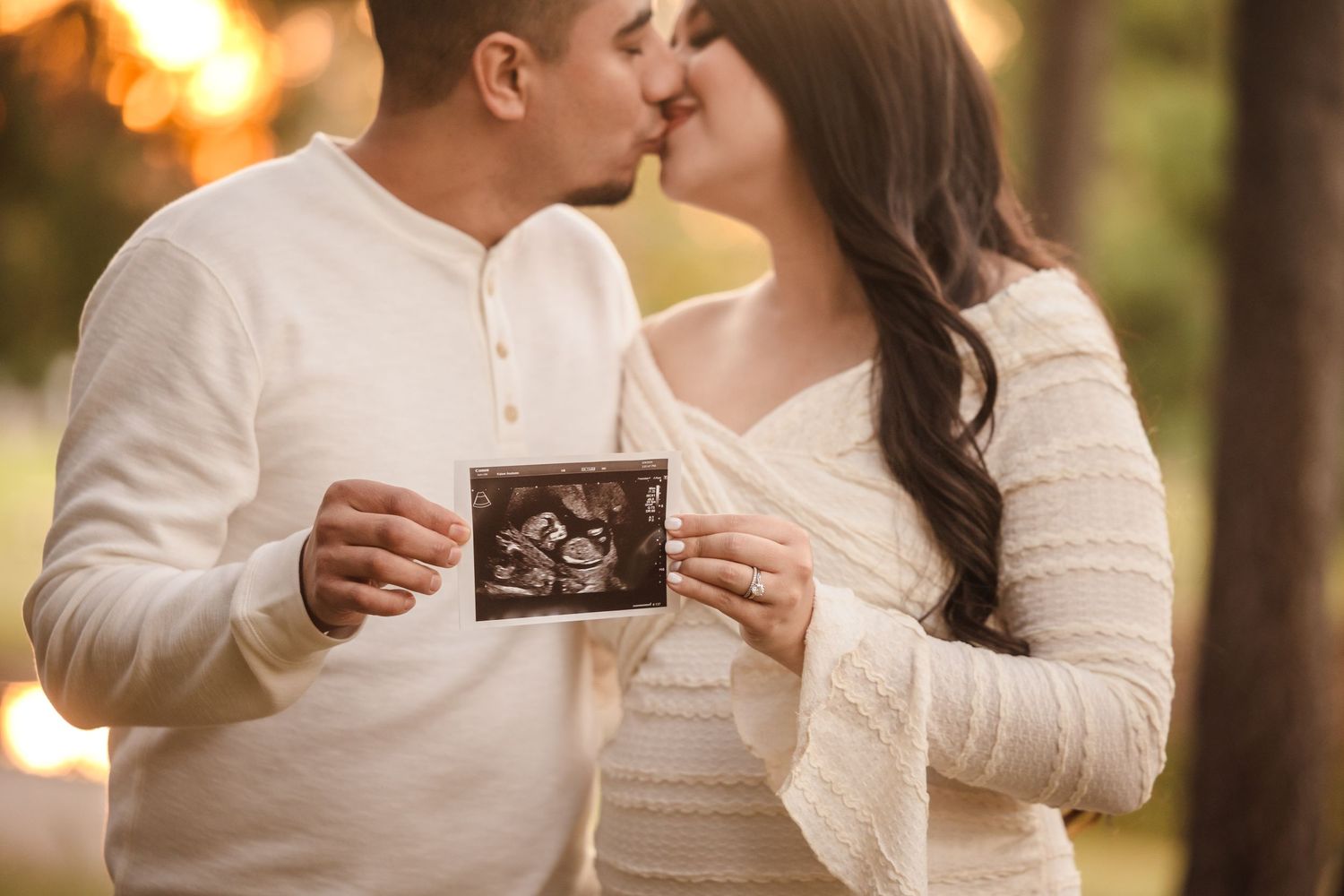 A couple in white sweaters share a kiss while holding an ultrasound photo during a pregnancy announcement photoshoot.