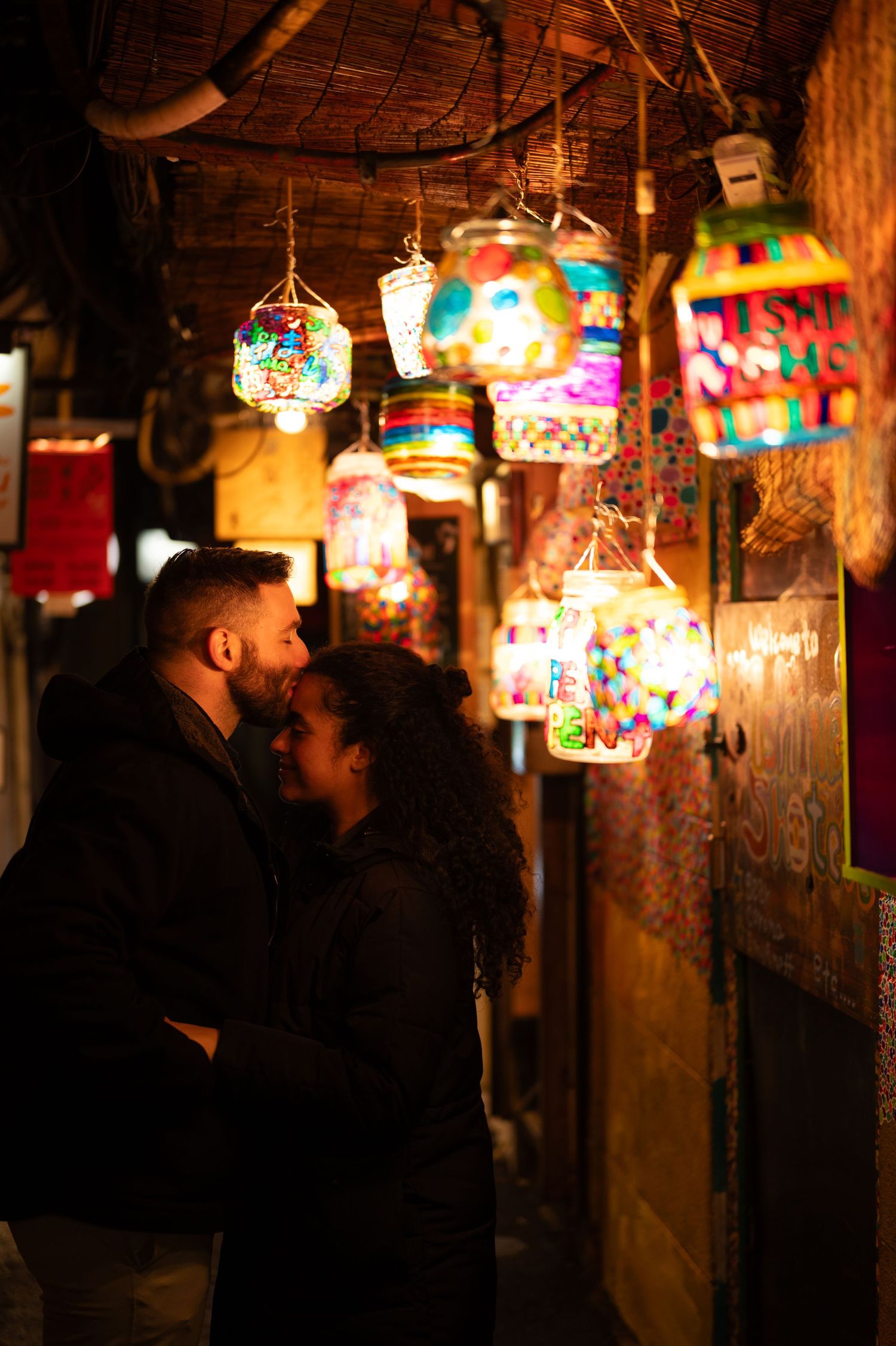 Romantic couple standing close together beneath colorful glowing lanterns in a vibrant market alley.