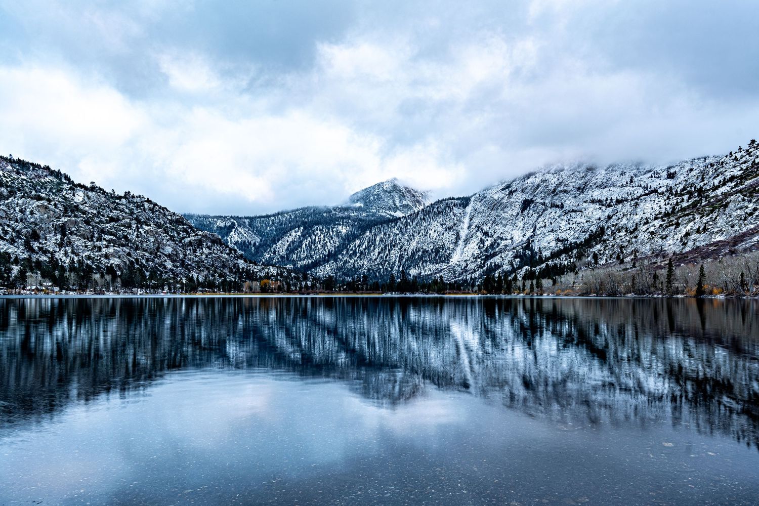 Snow-capped mountains reflect perfectly in still winter lake under cloudy sky.