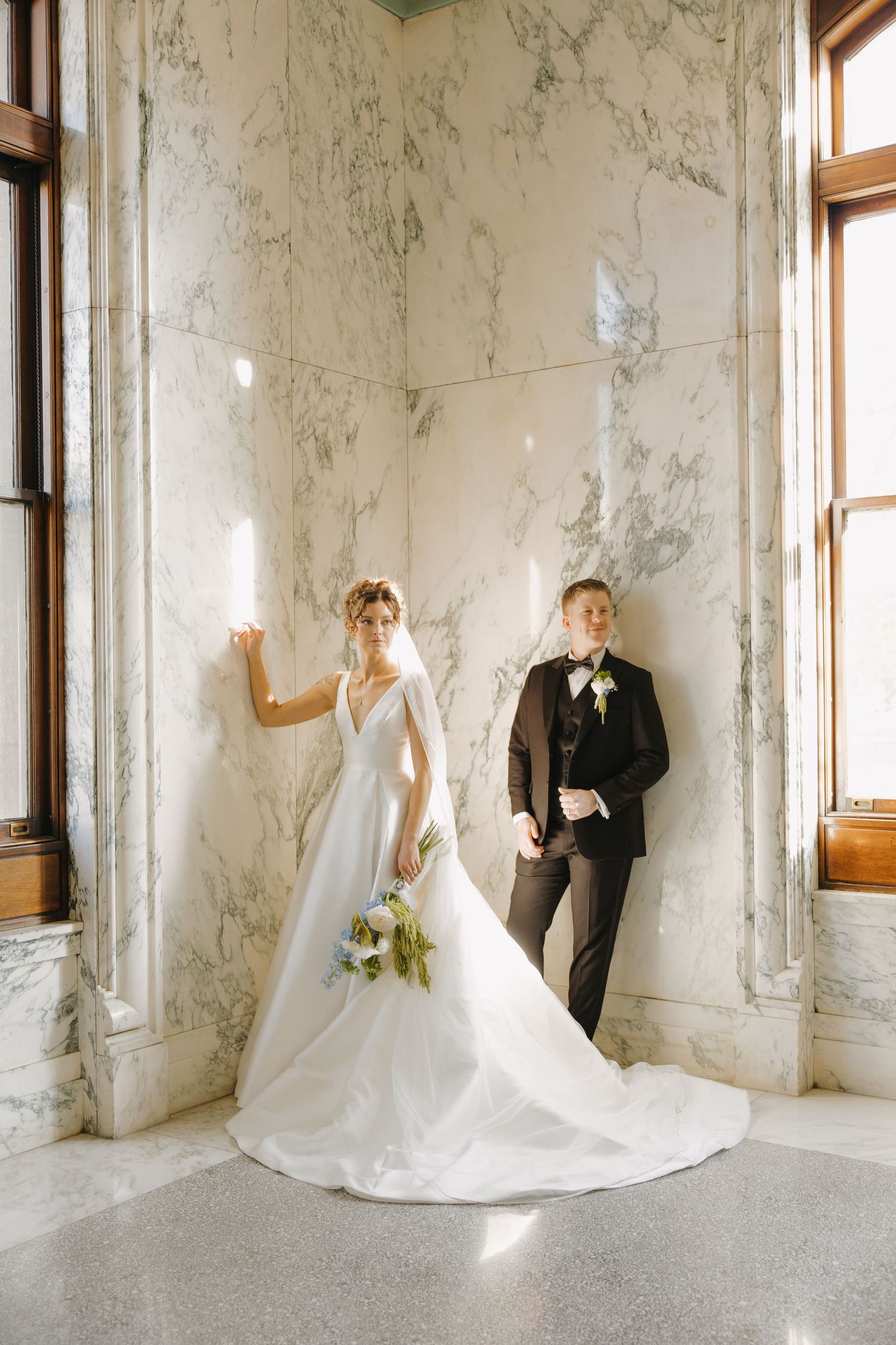 Bride in elegant ball gown and groom in black tuxedo posing in a marble-walled interior.