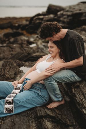 Couple lying on coastal rocks holding ultrasound photos together.