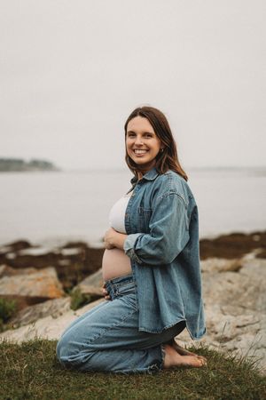 Expectant mother in denim sitting on coastal rocks by the ocean.