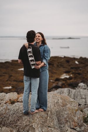 Happy couple holding ultrasound photos while standing on rocky coastline.