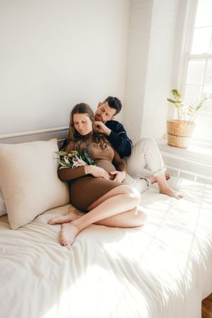 Intimate maternity photos showing couple relaxing together on white bedding in sunlit room.