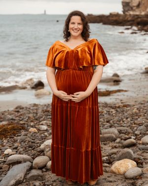 A maternity photoshoot on a rocky beach featuring a woman in a rust-colored velvet dress.