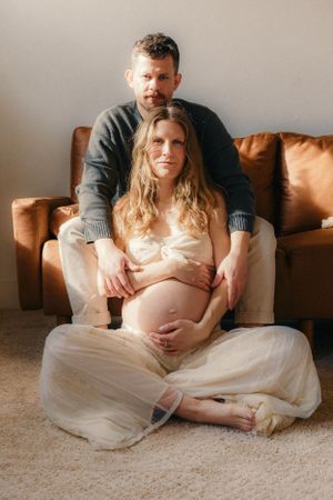 Maternity session showing couple sitting together on carpet with leather couch in background.