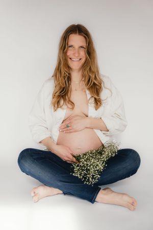Expectant mother in white shirt and jeans sits cross-legged with baby's breath flowers on white background.