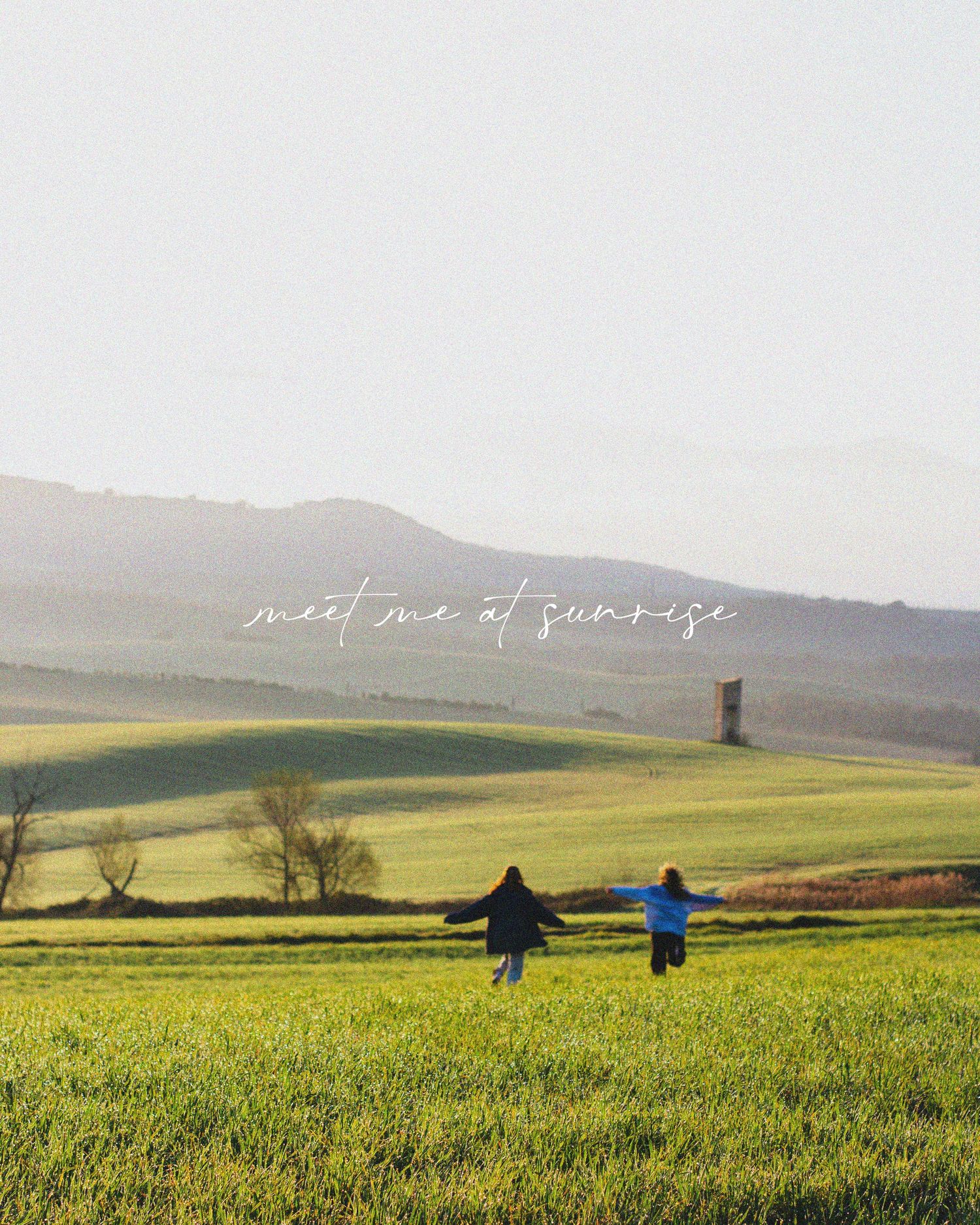 Two figures run joyfully through a green meadow at sunset with rolling hills and a lone structure in the misty background.