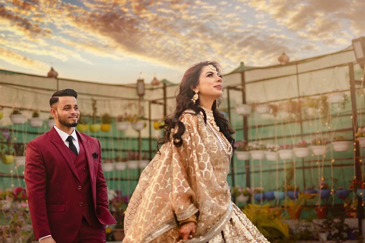 Two people in elegant formal attire pose outdoors against a sunset sky and glass building backdrop.
