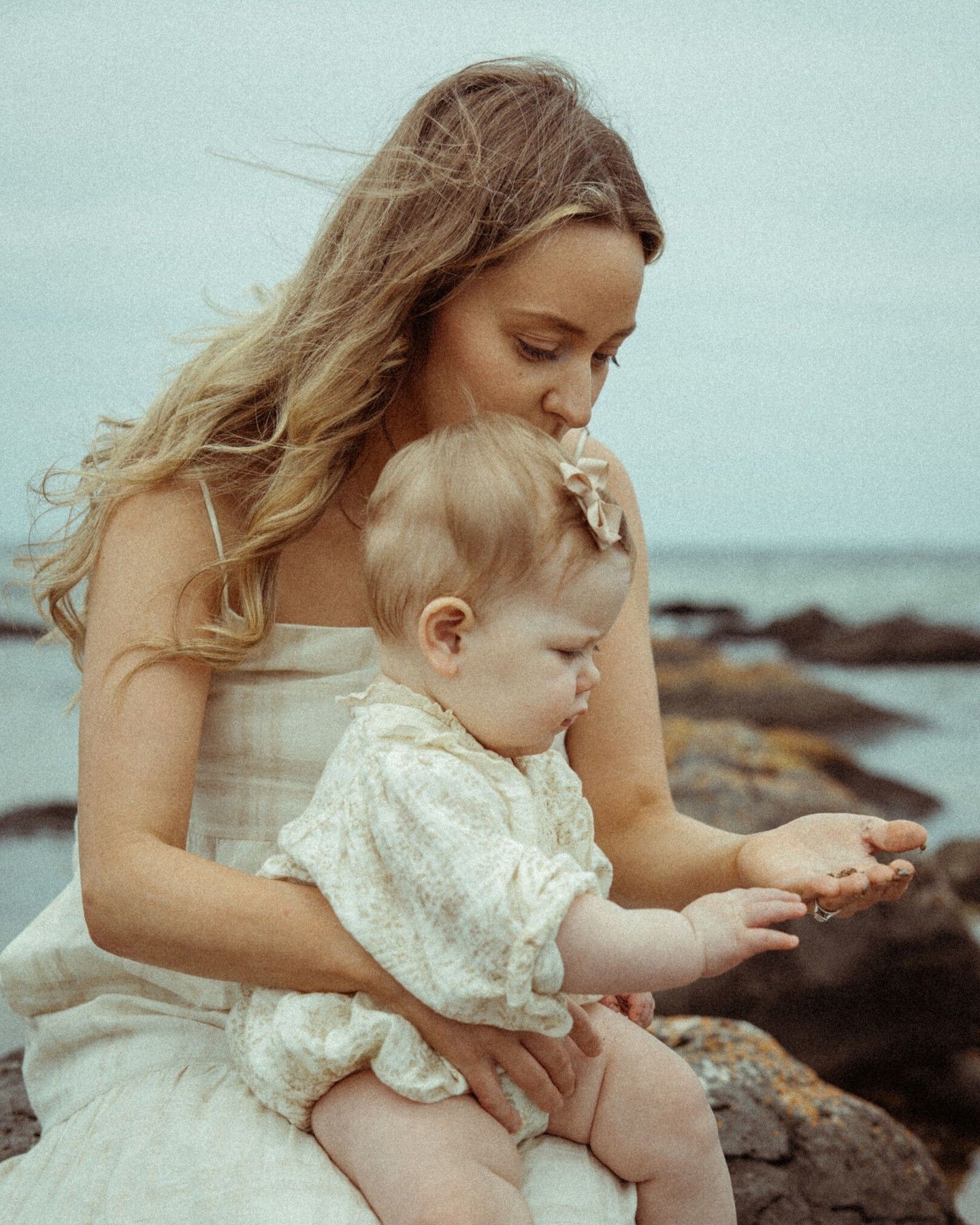 Mother and baby in white dresses sit together by the rocky oceanside on an overcast day.