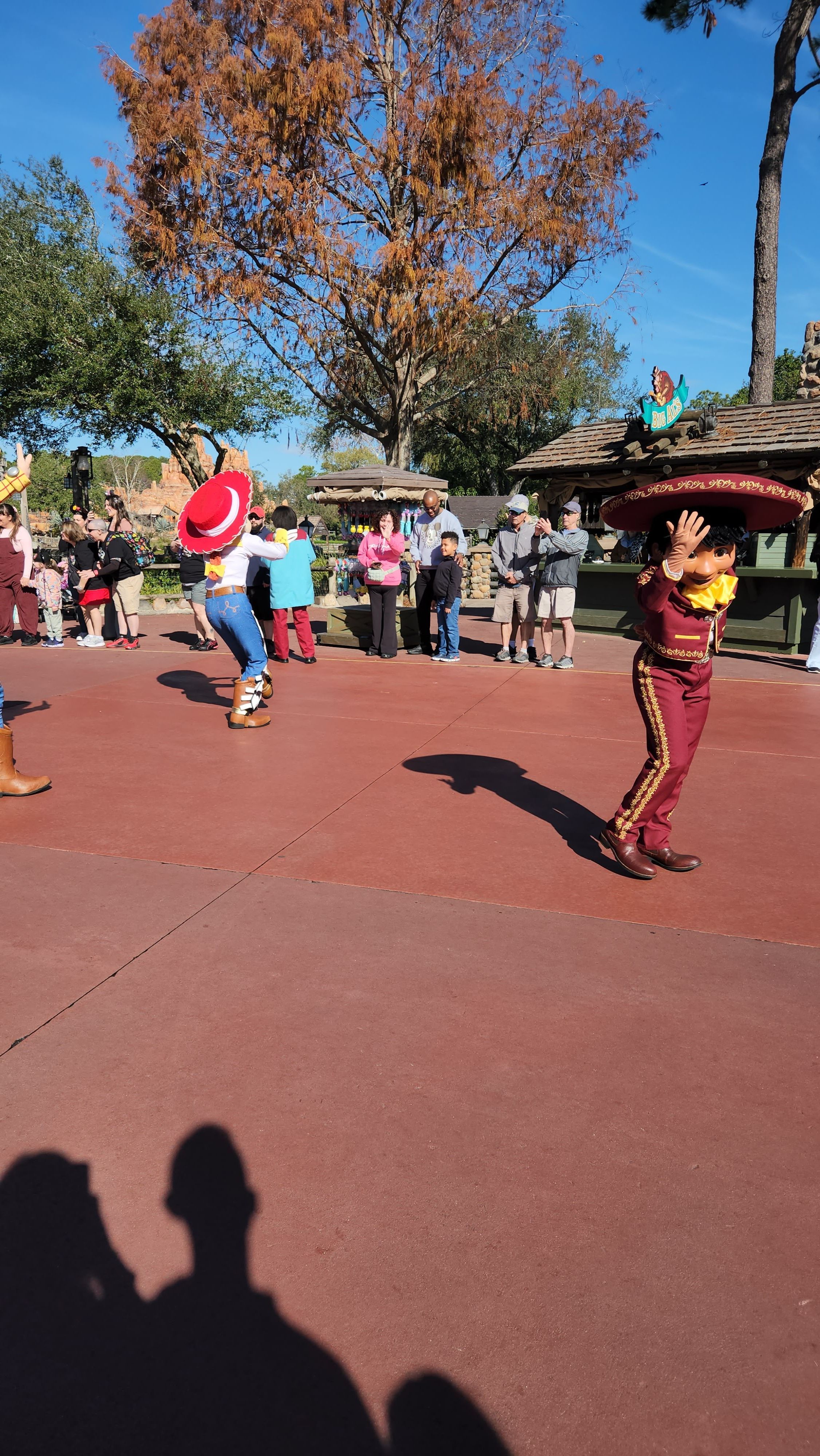 A crowd of Disney characters dancing and interacting with visitors on a red-paved outdoor area during a sunny autumn day.