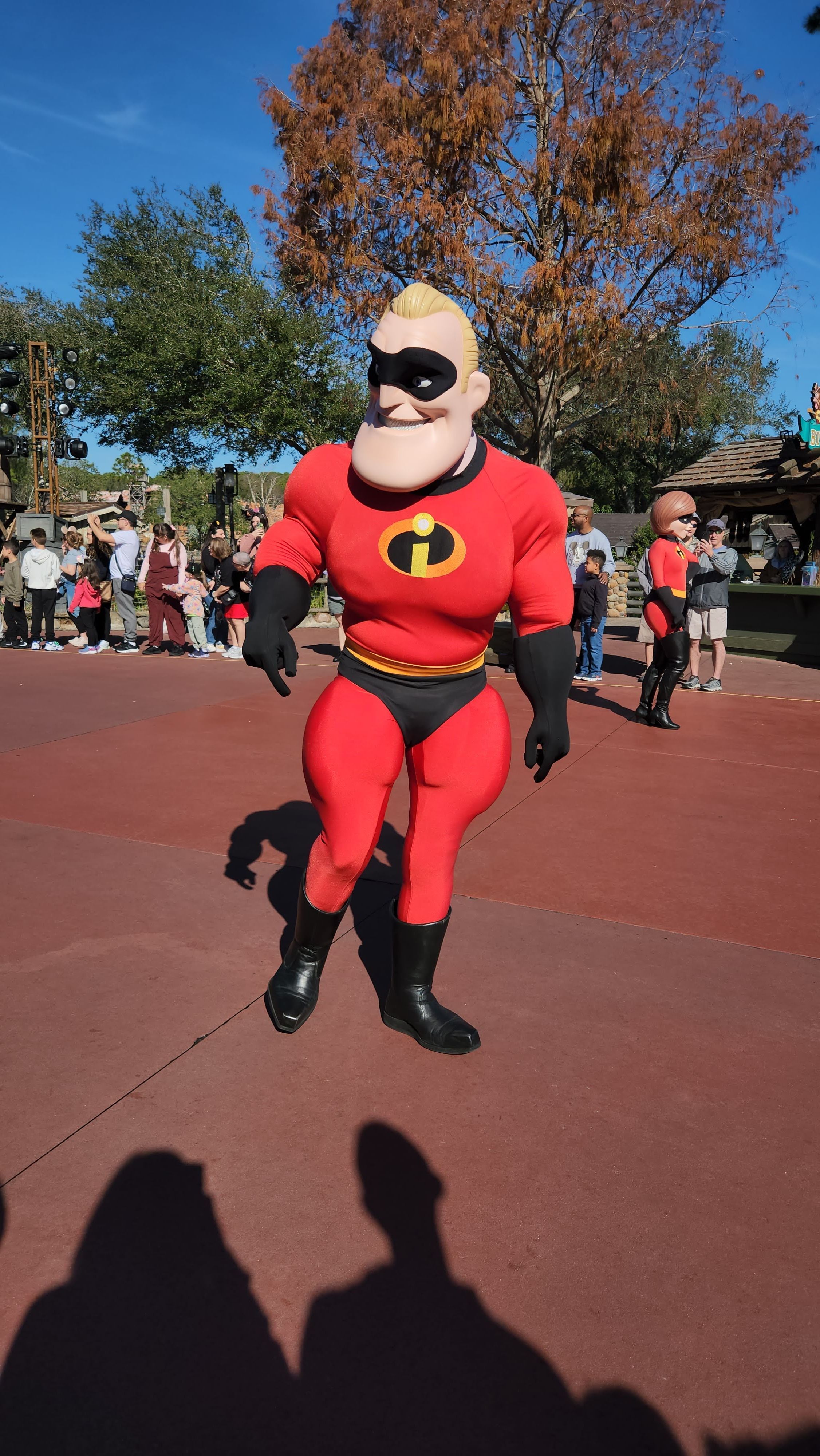 Mr. Incredible character in costume poses on red pavement at a Disney theme park with autumn trees in background.