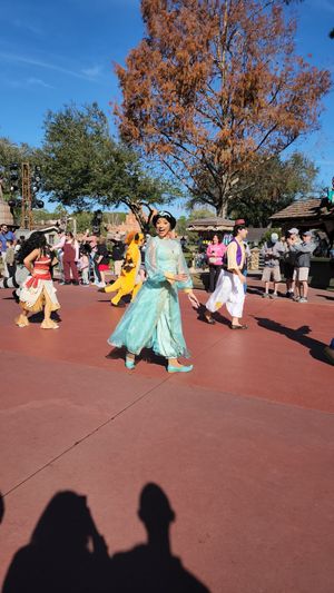 Costumed performers in flowing dresses dance and twirl on a red pavement plaza during a theme park show performance.