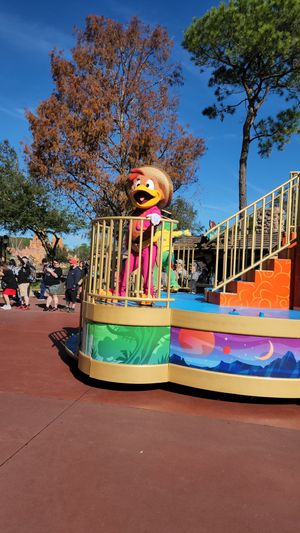 Colorful parade float with yellow puppet character on display at theme park under blue sky with autumn trees.