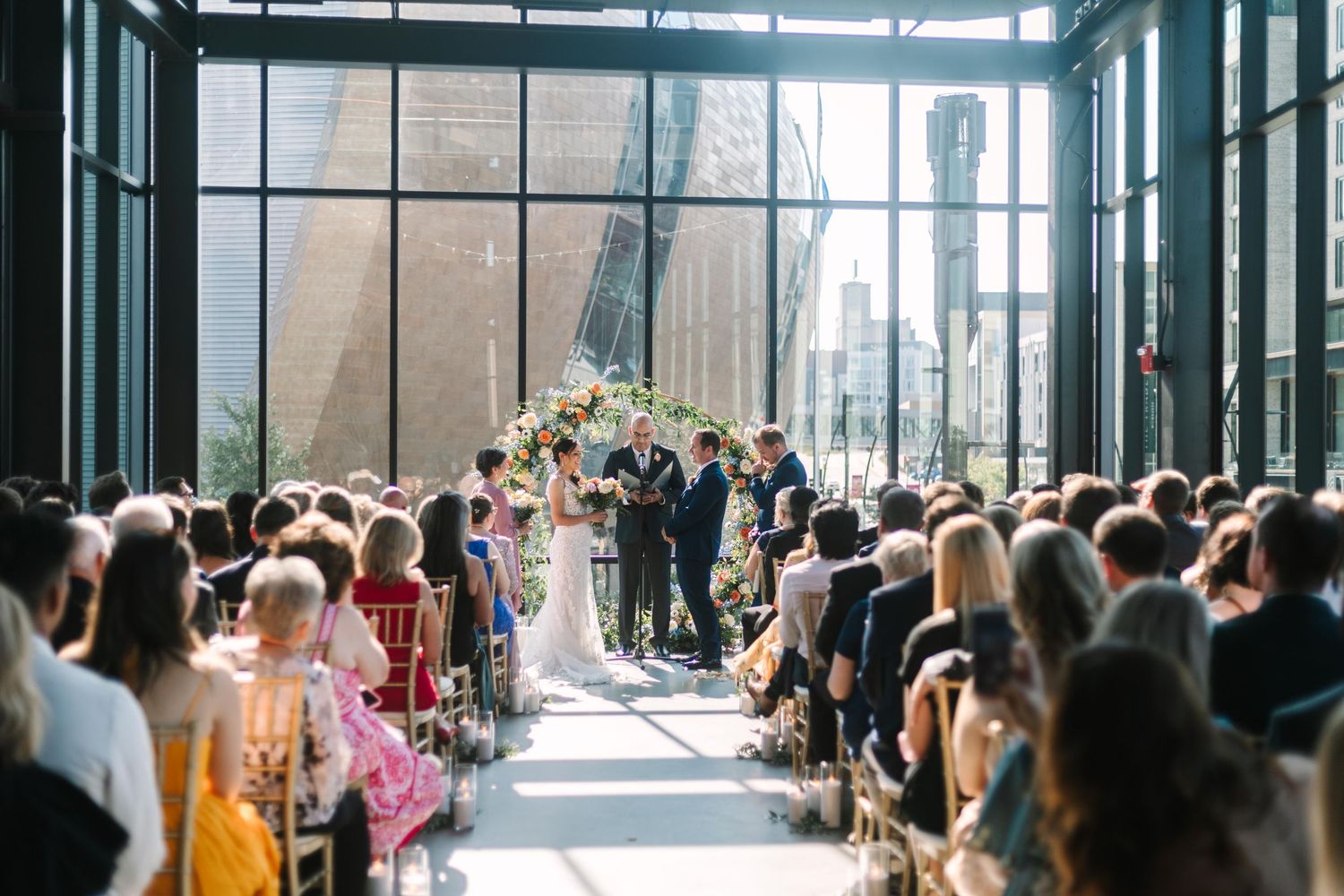 A wedding ceremony takes place in a modern glass-walled venue with guests seated in rows as natural light streams in.