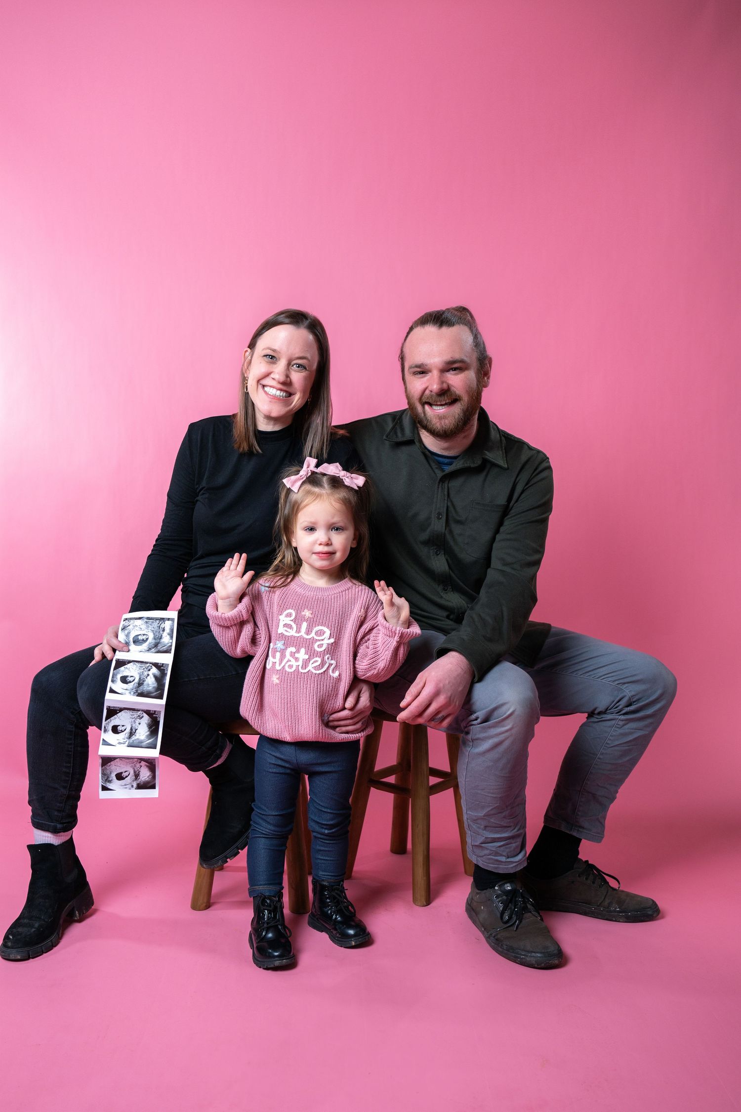A family poses together against a pink backdrop with ultrasound photos displayed during a pregnancy announcement photoshoot.