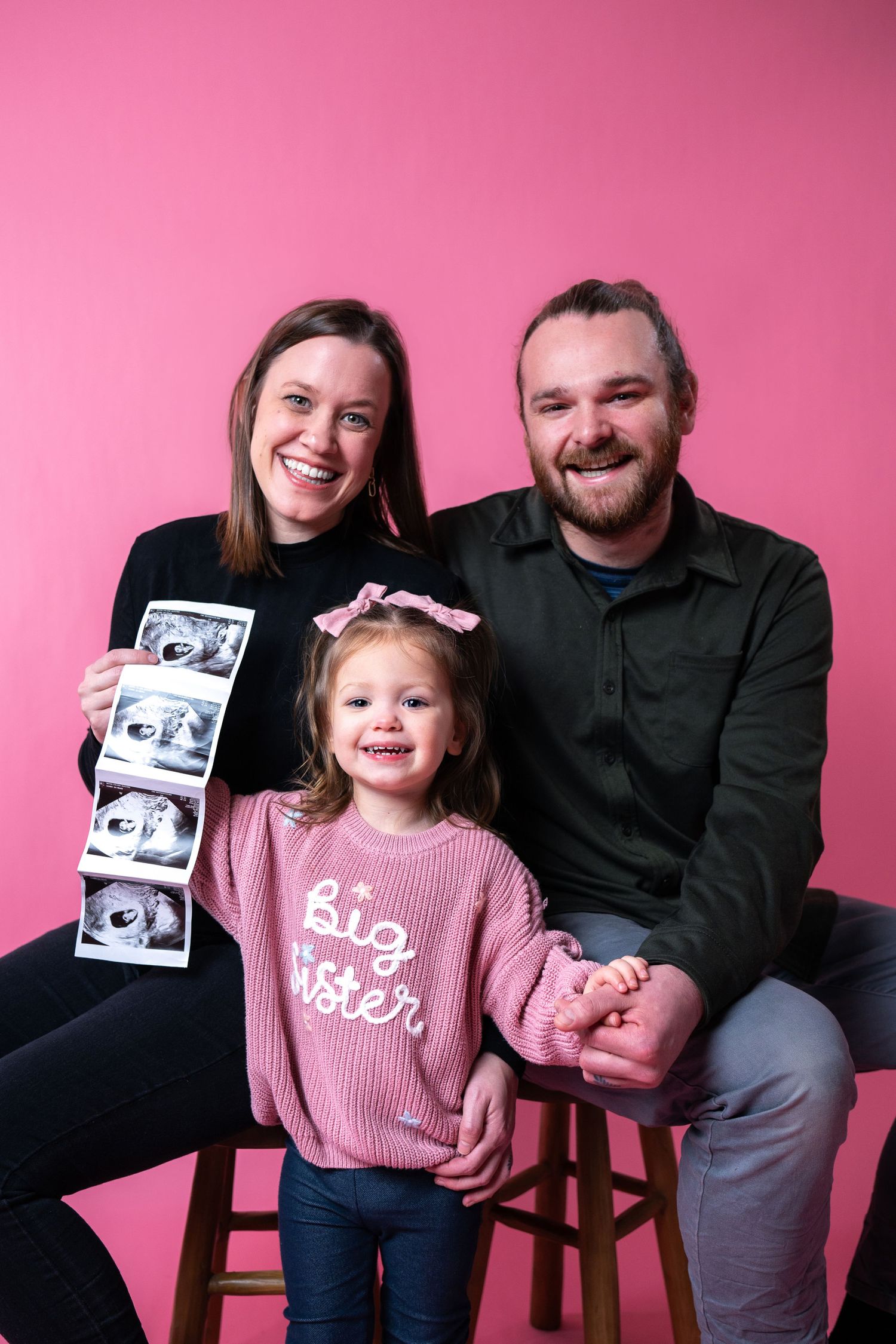 A family poses against a pink backdrop while holding ultrasound photos and wearing a 'big sister' sweater.