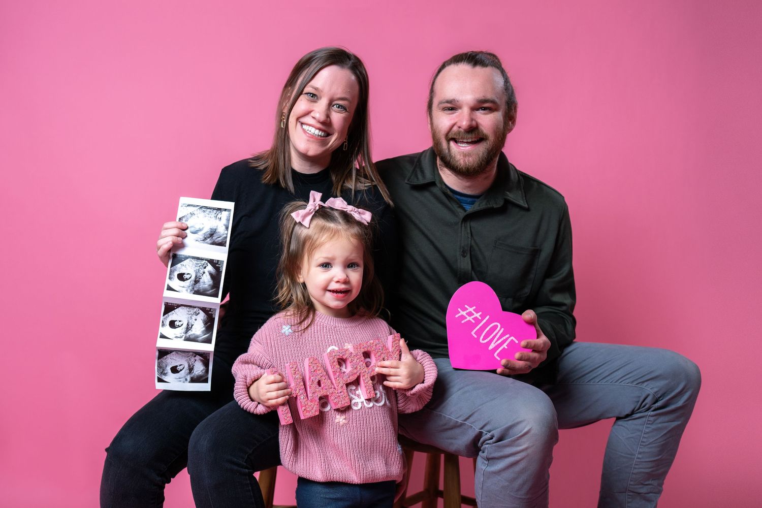 A family poses together against a pink backdrop holding ultrasound photos and a heart sign.