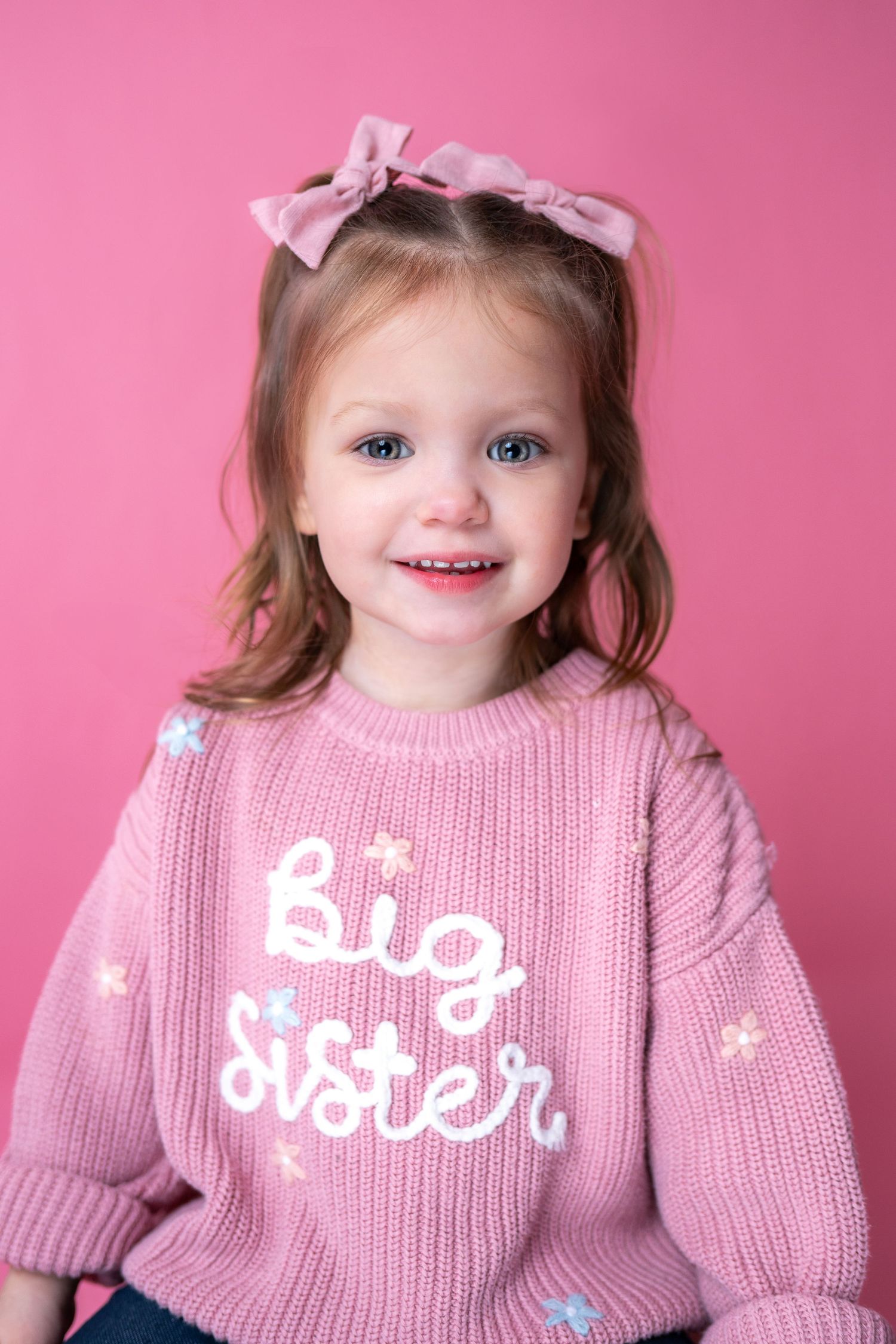 A child in a pink 'Big Sister' sweater smiles while wearing hair bows against a pink background.