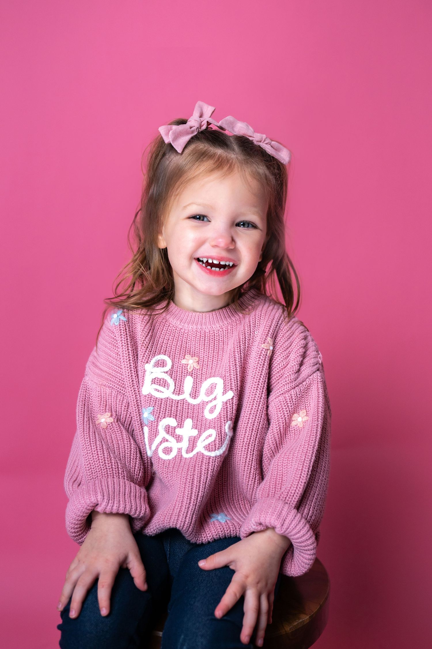 A young child wearing a pink 'Big Sister' sweater sits and smiles against a bright pink background.
